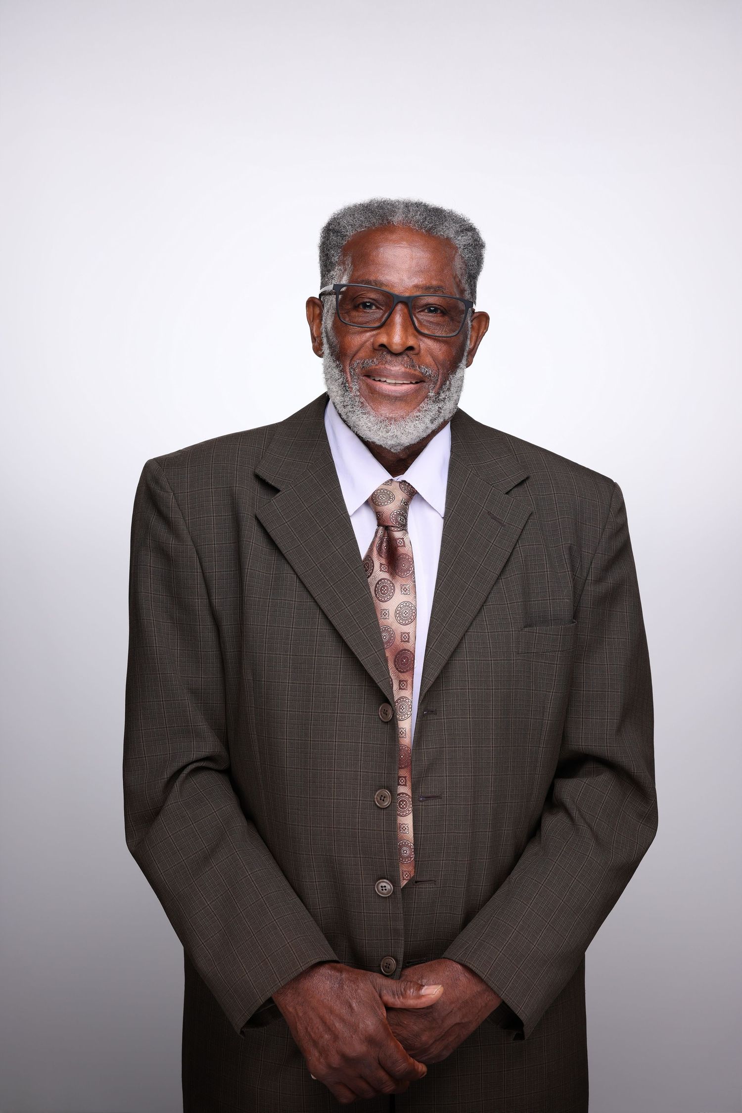 Elderly Black man in brown suit, tie, and glasses smiling warmly against a light gray background.