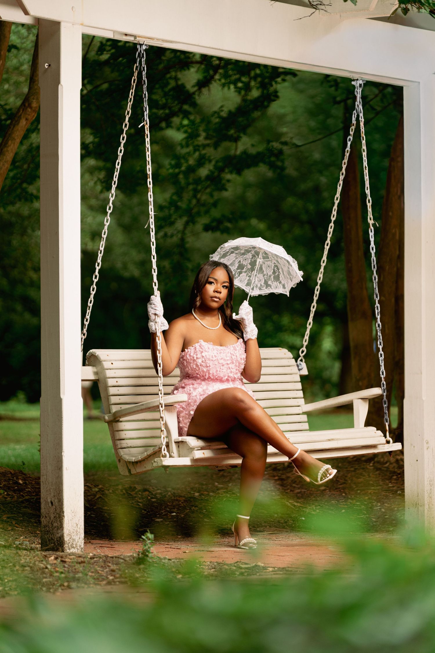 Young Black woman in pink mini dress holding lace umbrella, seated on white wooden porch swing surrounded by lush greenery.