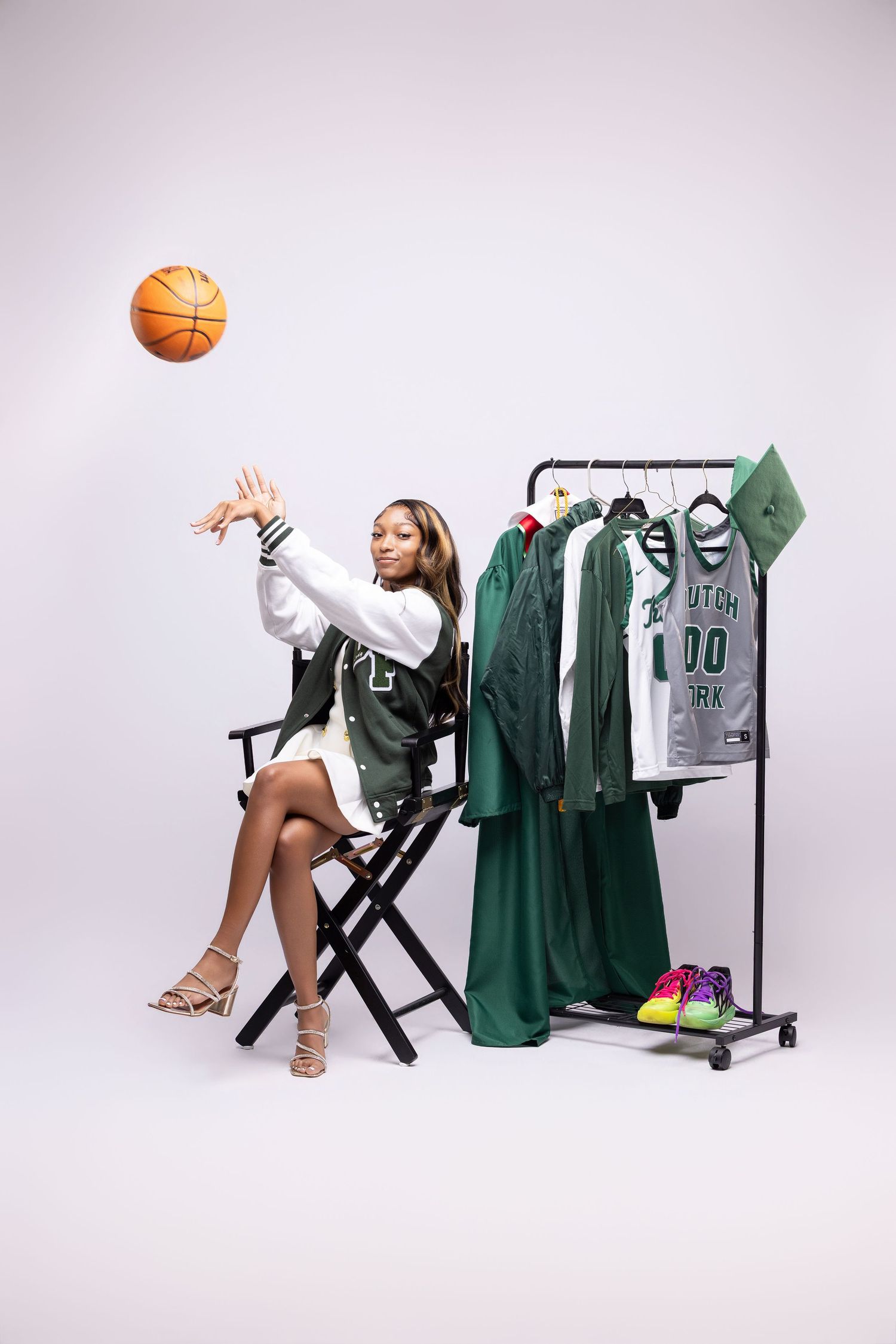 Woman in green varsity jacket tosses basketball beside a clothing rack with Dutch Fork High School jerseys in a studio setting.