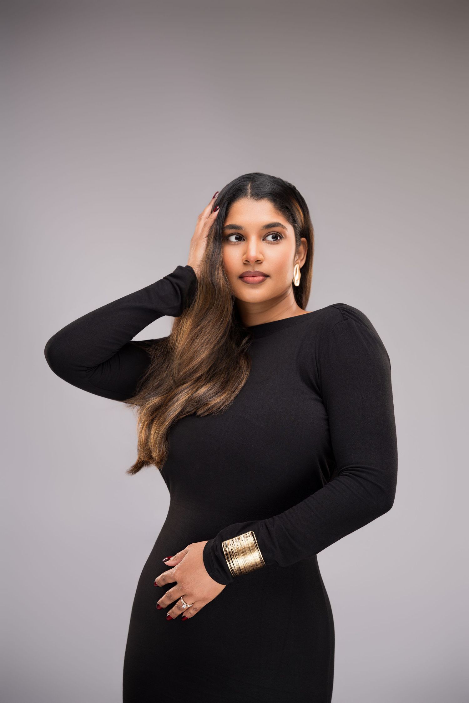 Woman in black long-sleeve dress with gold bangles and earrings, posing gracefully against a gray background.