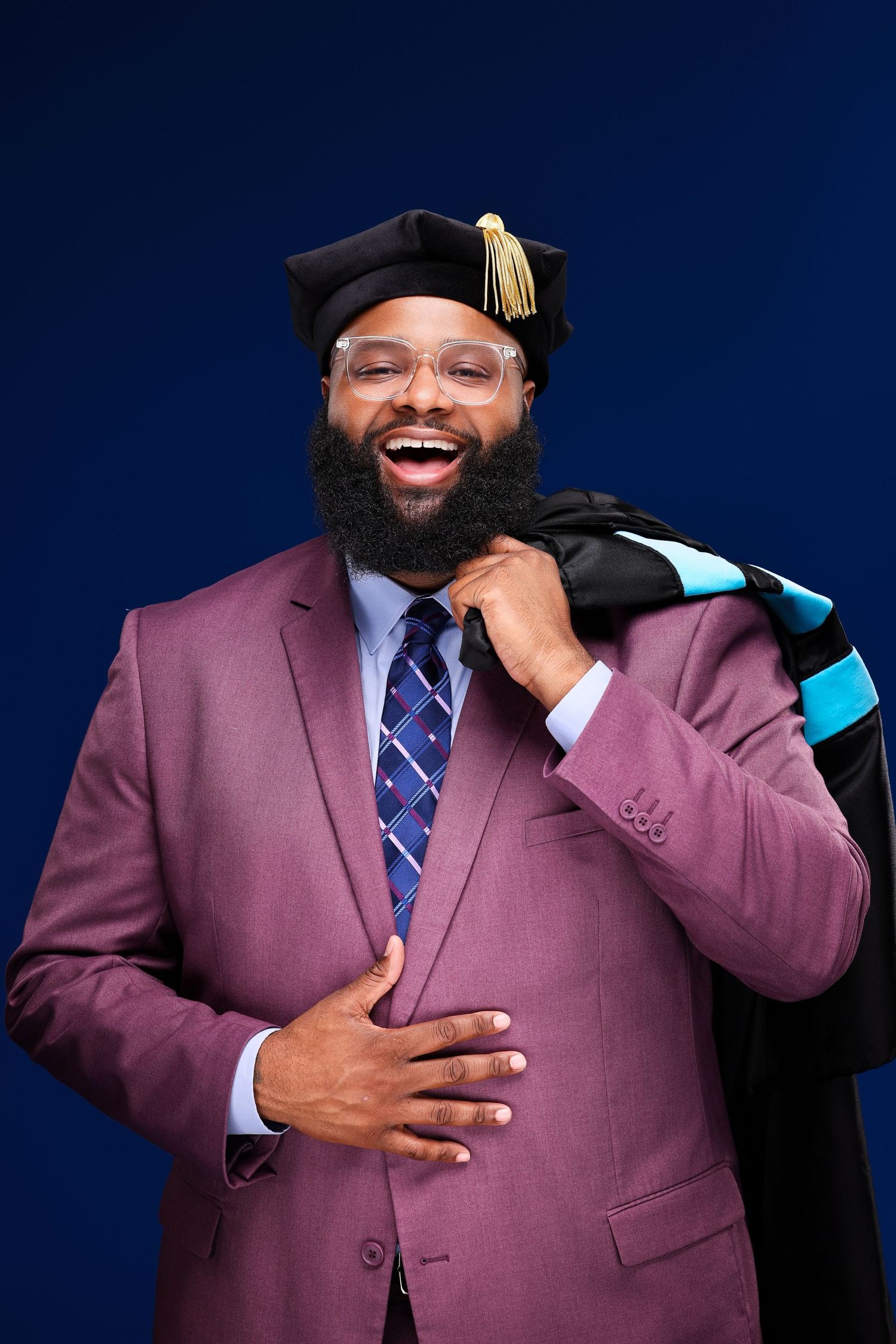 A graduate in a purple suit and graduation cap laughs joyfully while holding their chest during a photoshoot.