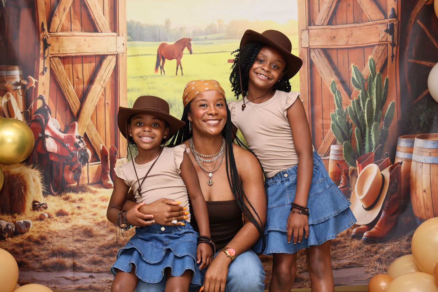 A woman and two children pose in cowboy hats and denim skirts in front of a Western barn backdrop.