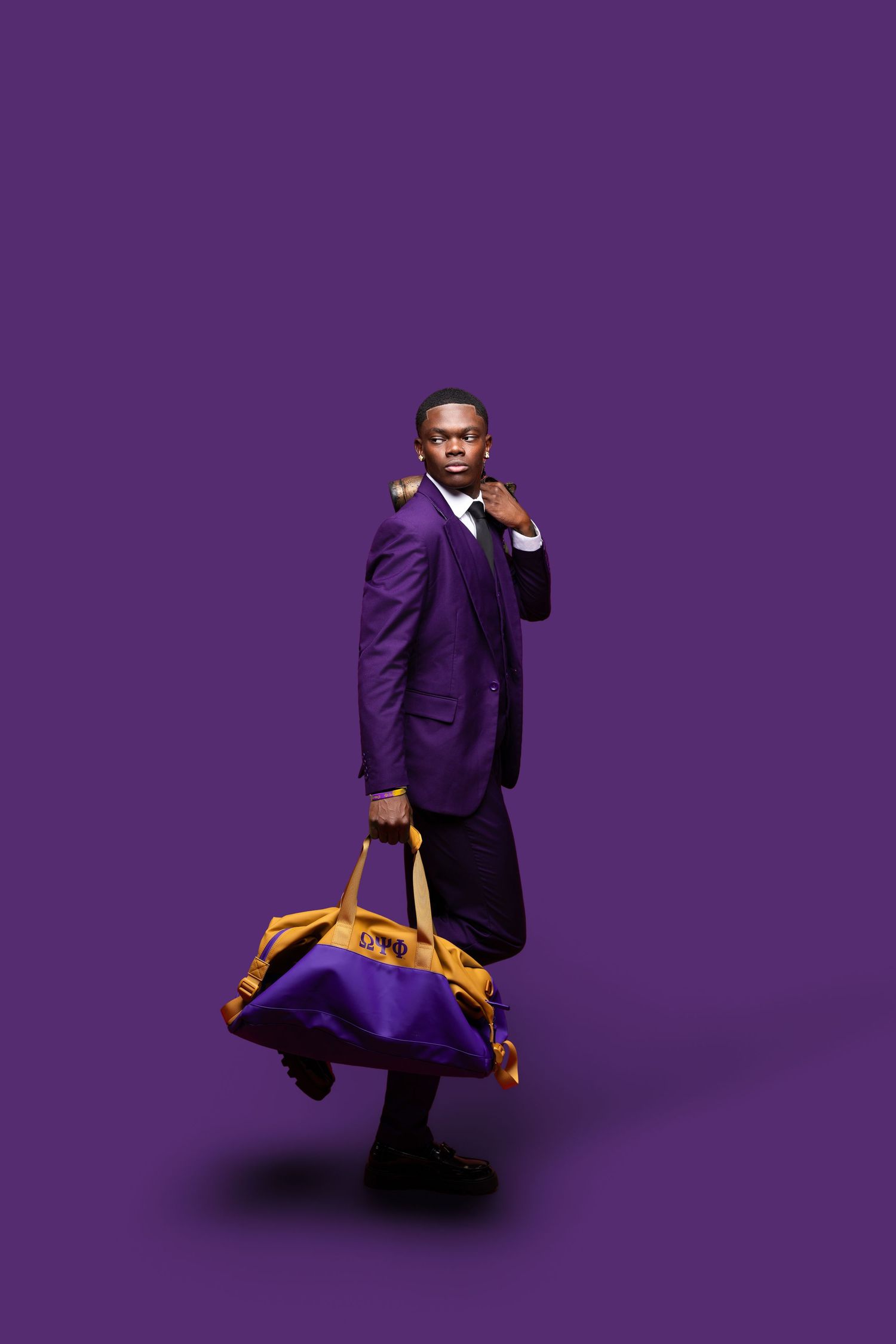 A stylish man in a purple suit carries a duffel bag against a purple background.