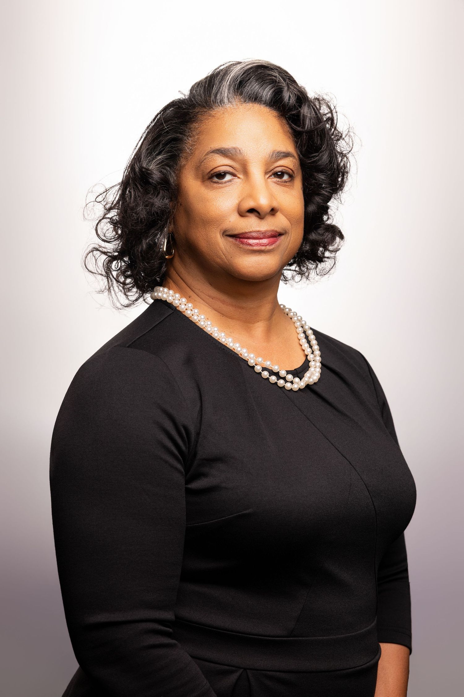 Professional woman in black dress and pearl necklace poses for formal portrait against light gray background.