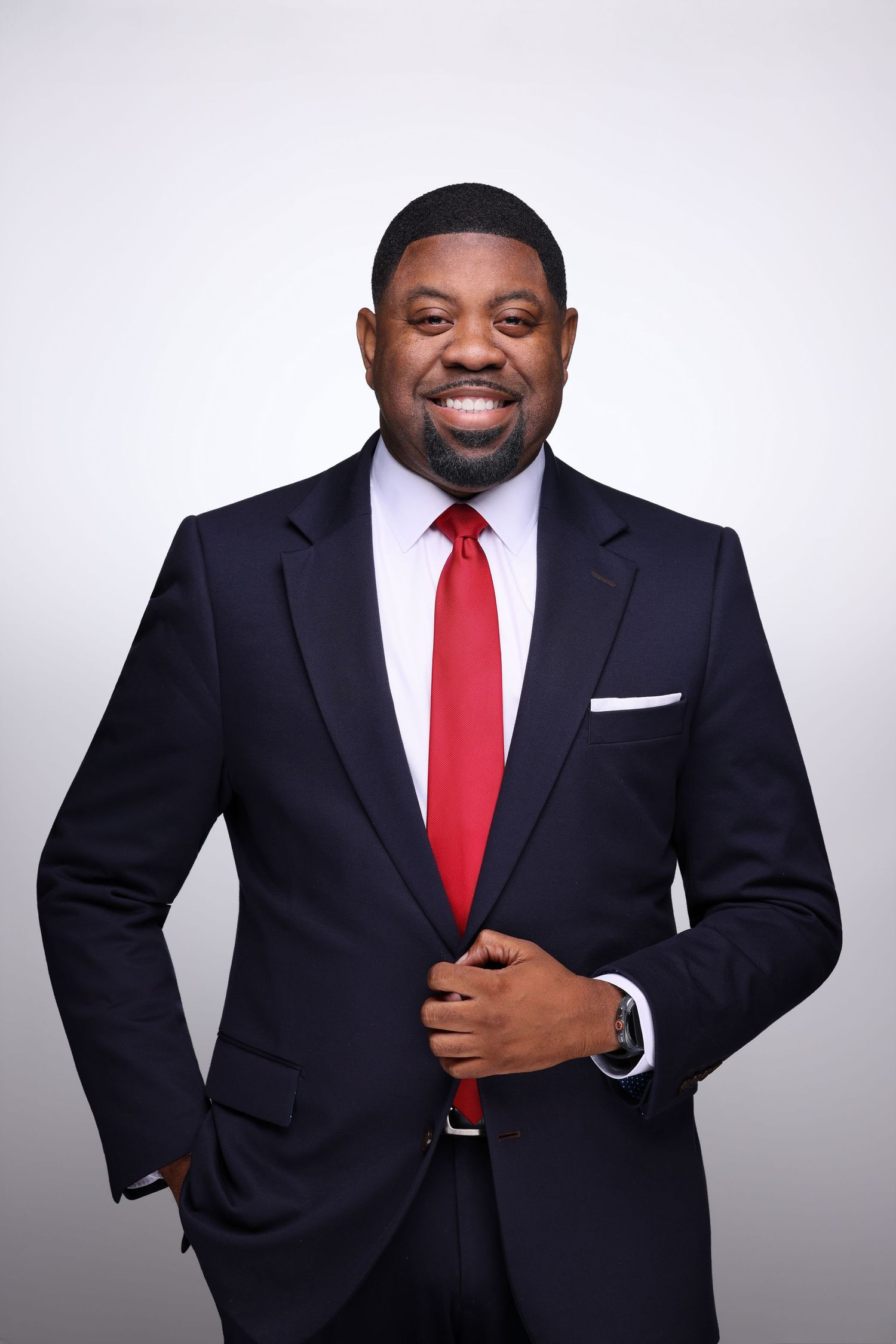 Smiling professional man in dark suit with red tie posing confidently against a gray background.