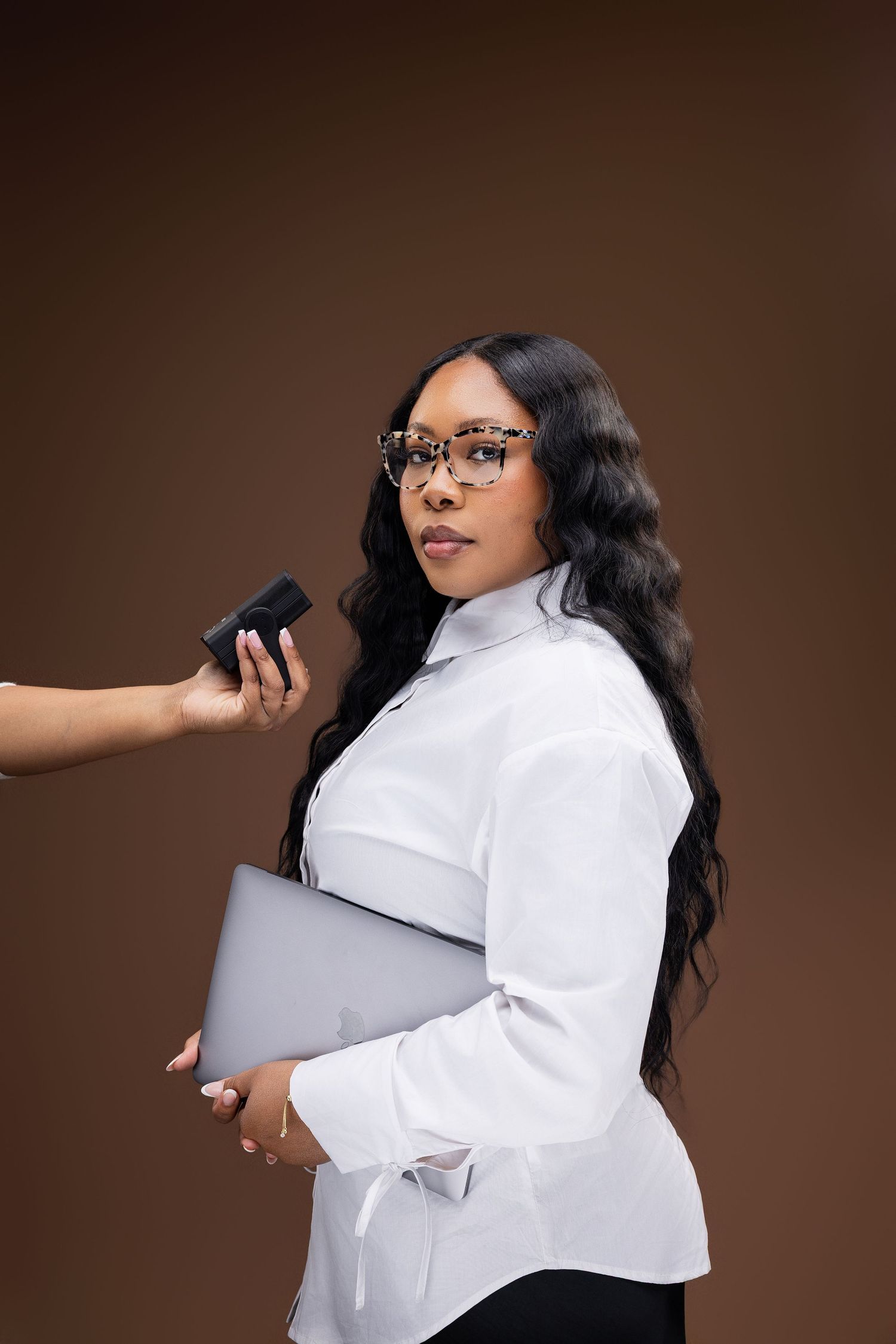 Professional wearing glasses and a crisp white button-down shirt holds a laptop while someone applies makeup in a studio setting.