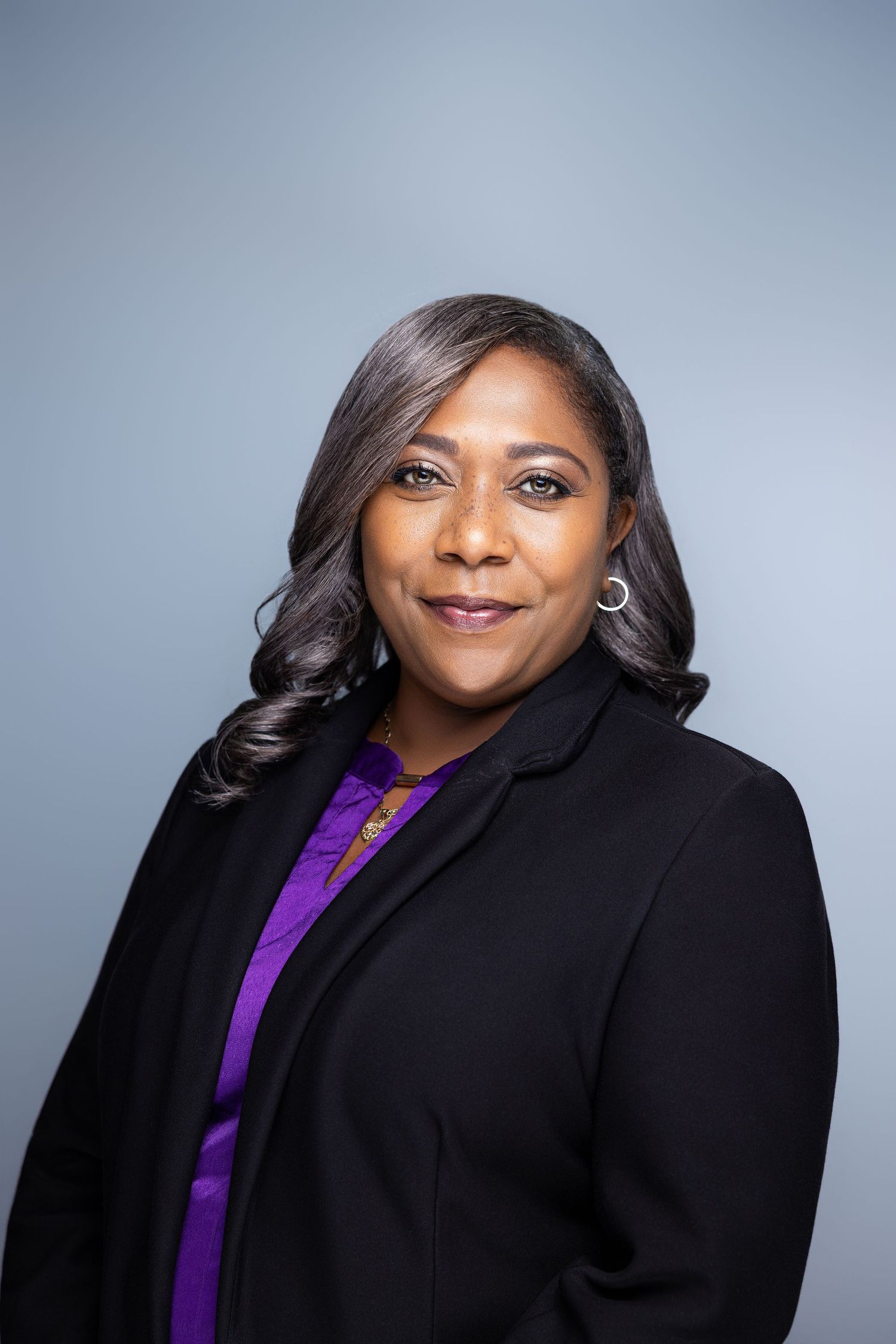 Professional woman in black blazer and purple blouse smiling confidently against a gray background.