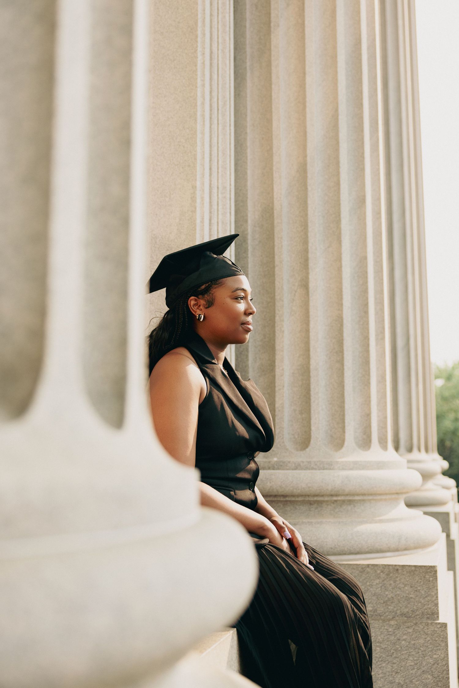 Graduate in cap and gown sitting thoughtfully beside classical stone columns.