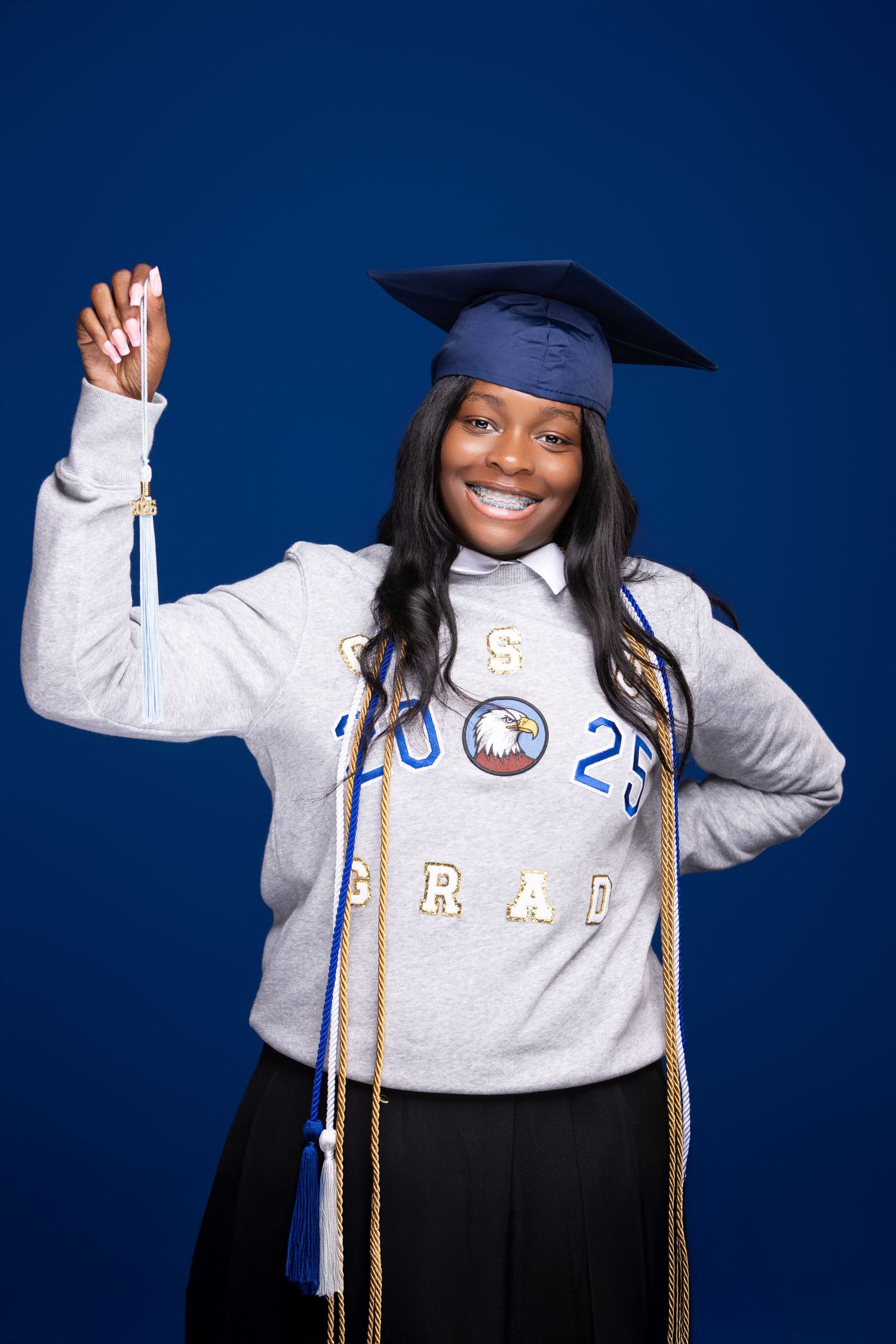 Graduate in blue cap and grey sweatshirt celebrating with raised arm against blue background.