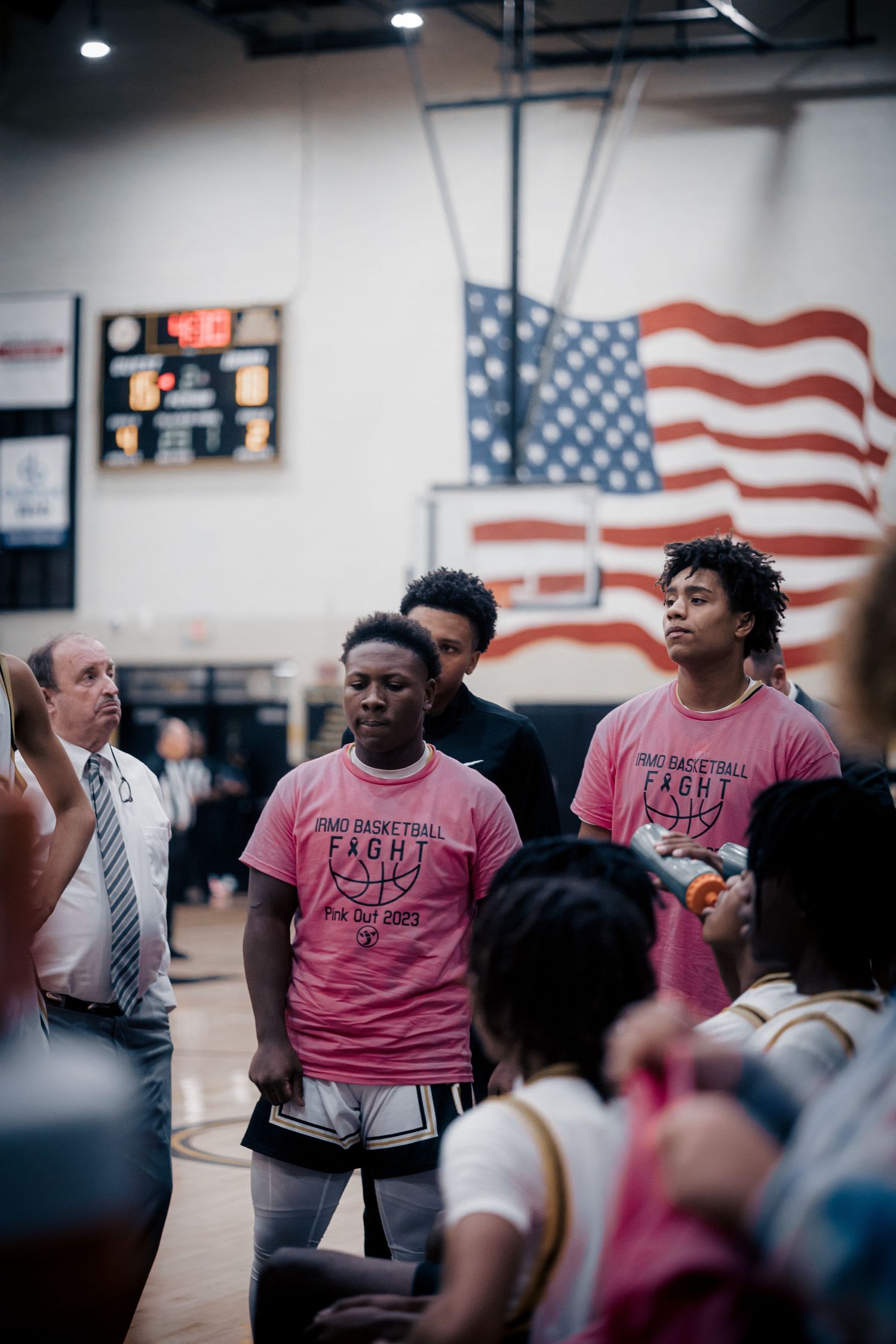 People gathered in gymnasium with American flag display and scoreboard visible in background.
