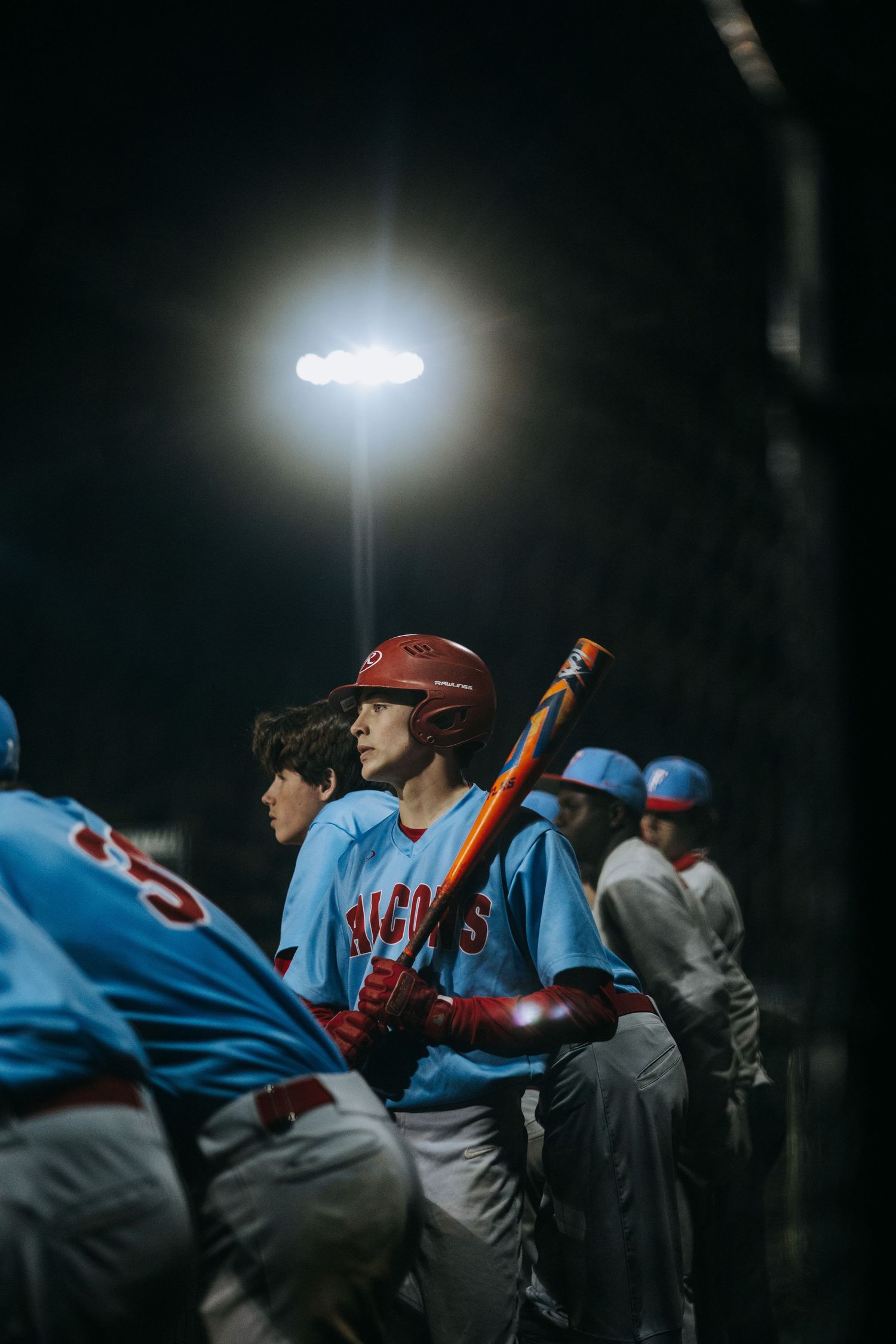 Baseball players in light blue uniforms stand in dugout under bright stadium lights.