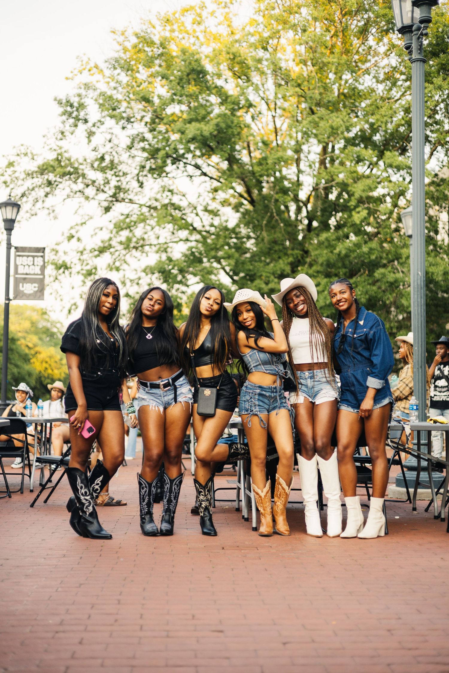 Group of friends in denim shorts and cowboy boots pose together on brick patio on sunny autumn day.