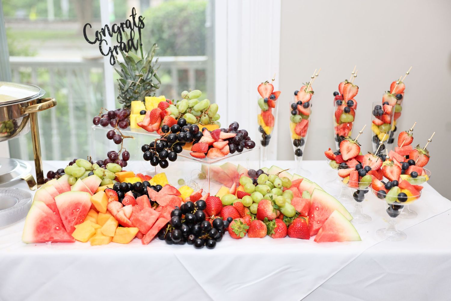 Colorful graduation fruit display with watermelon, grapes, pineapple, and fruit skewers on a white table.