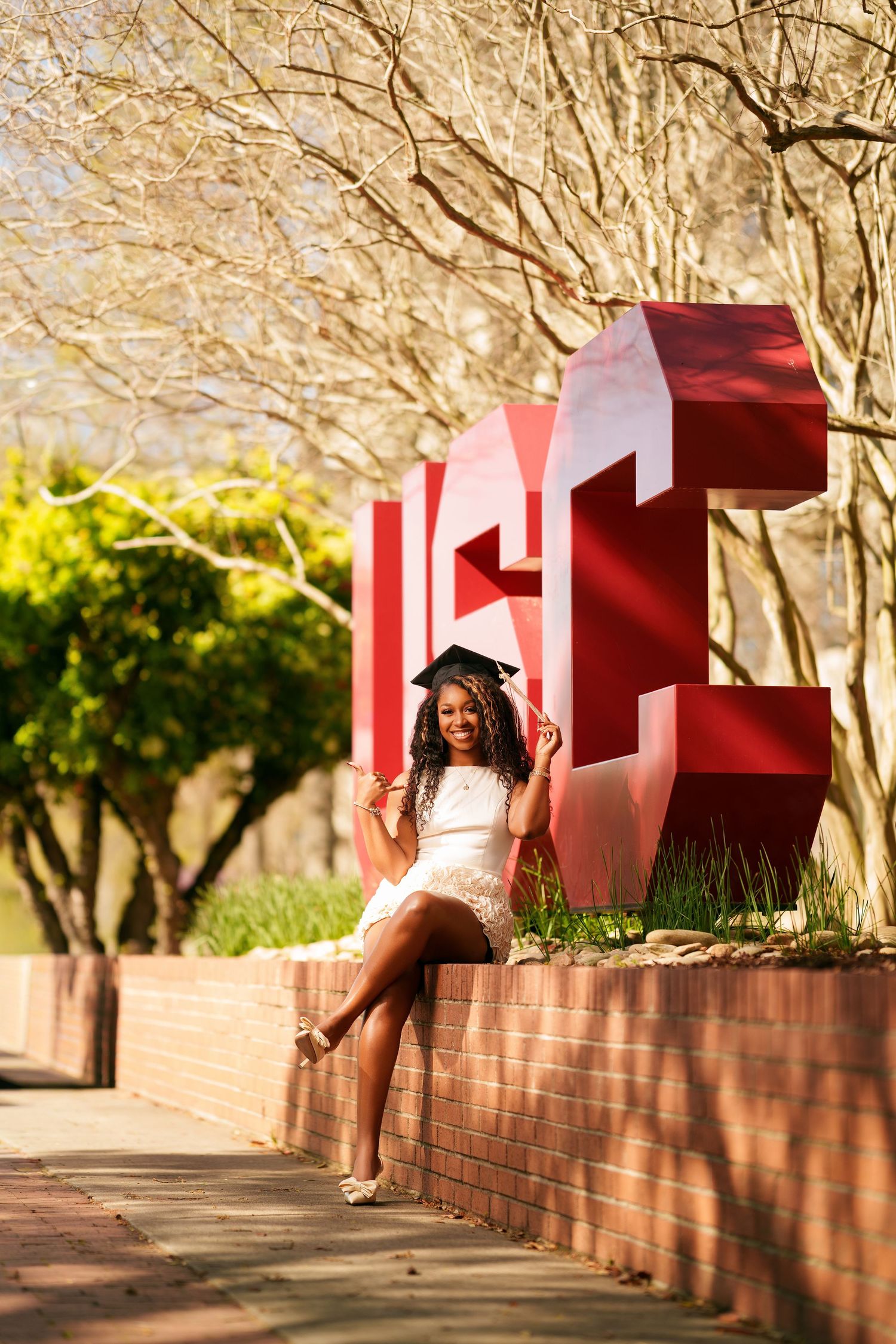 Young woman in white dress and graduation cap poses proudly beside large red university letters on a sunny campus.