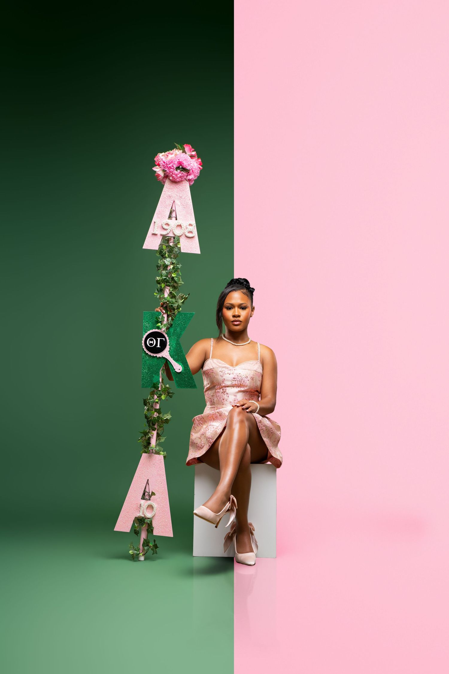 Woman in pink dress seated beside Greek sorority letters on green and pink split background.