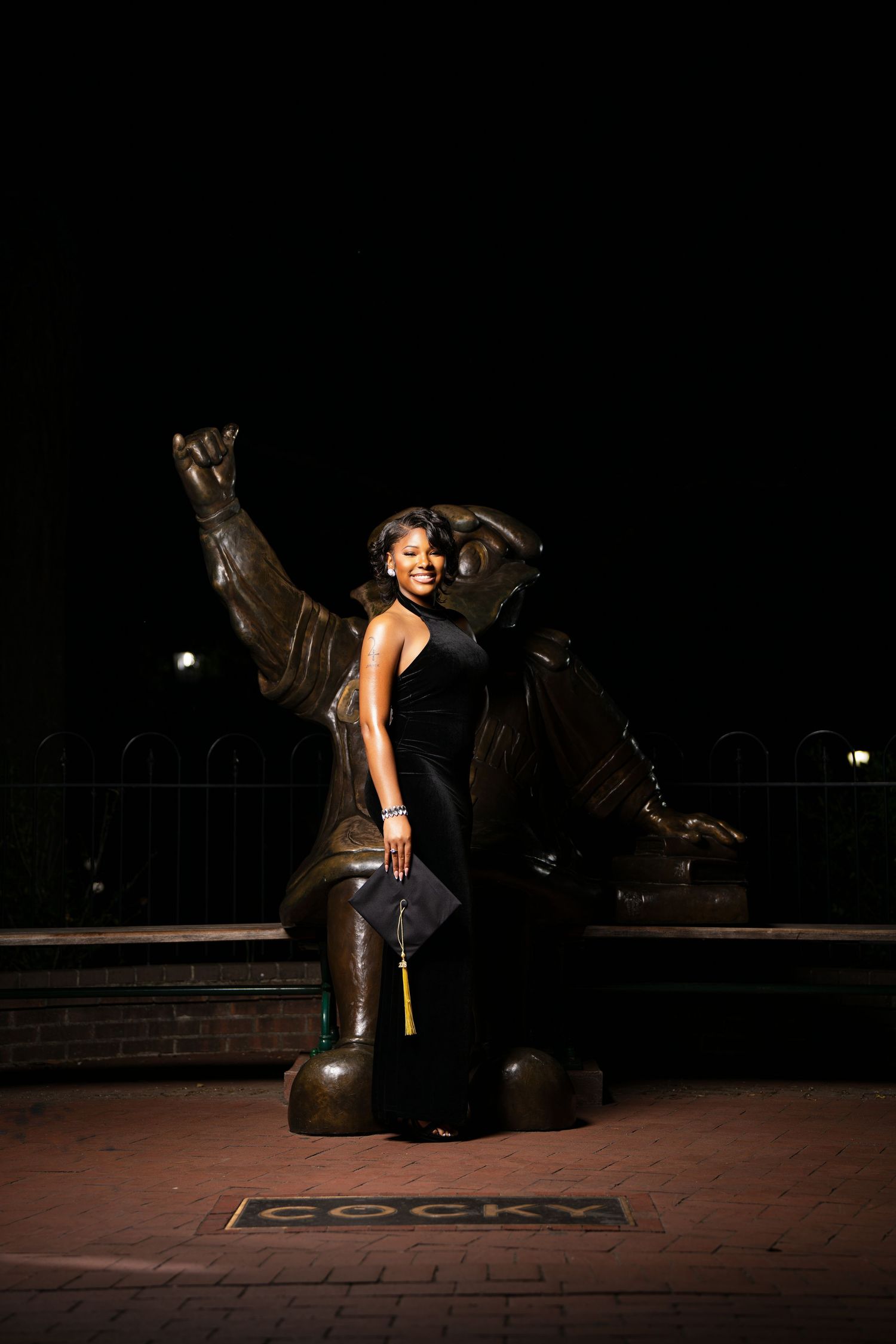 Graduate in black gown poses proudly before the Cocky mascot statue at night, holding her graduation cap.