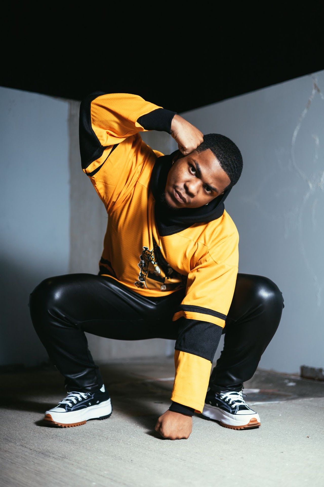 Athletic person in yellow sweatshirt and black pants crouches in dynamic pose wearing classic sneakers against gray wall.