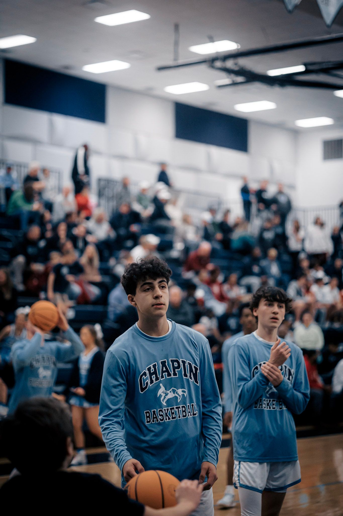 Students wearing Chapin basketball spirit wear watch game from crowded gymnasium.