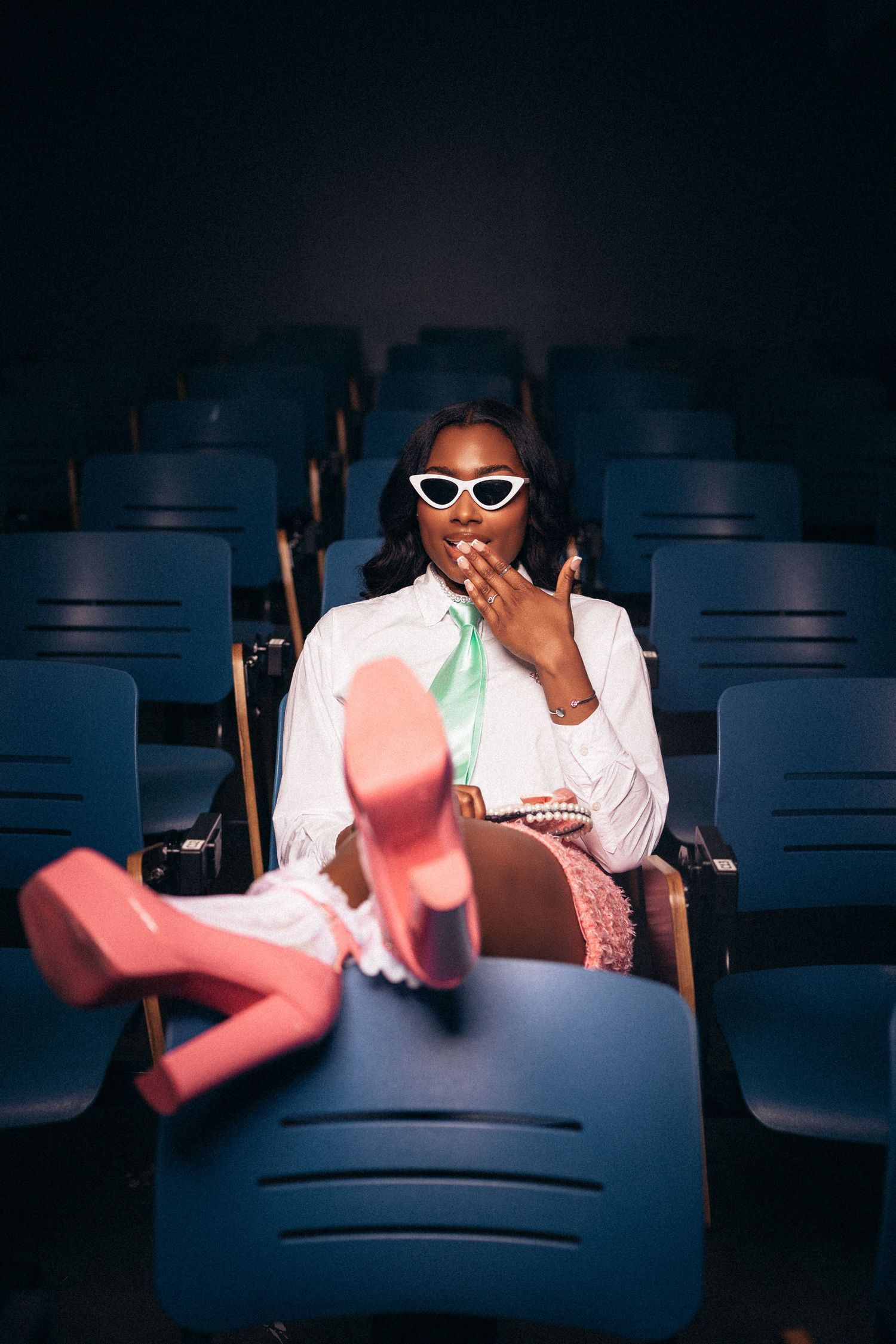 Person in white outfit and sunglasses sitting in dark theater with pink lighting accents.