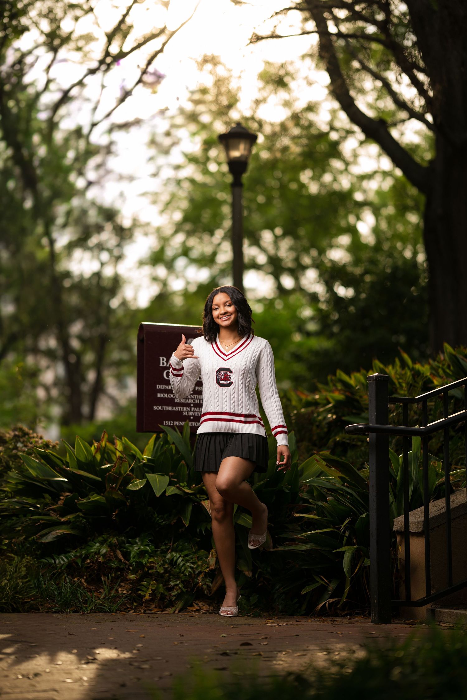 Young woman in University of South Carolina sweater giving thumbs up on a tree-lined campus path.