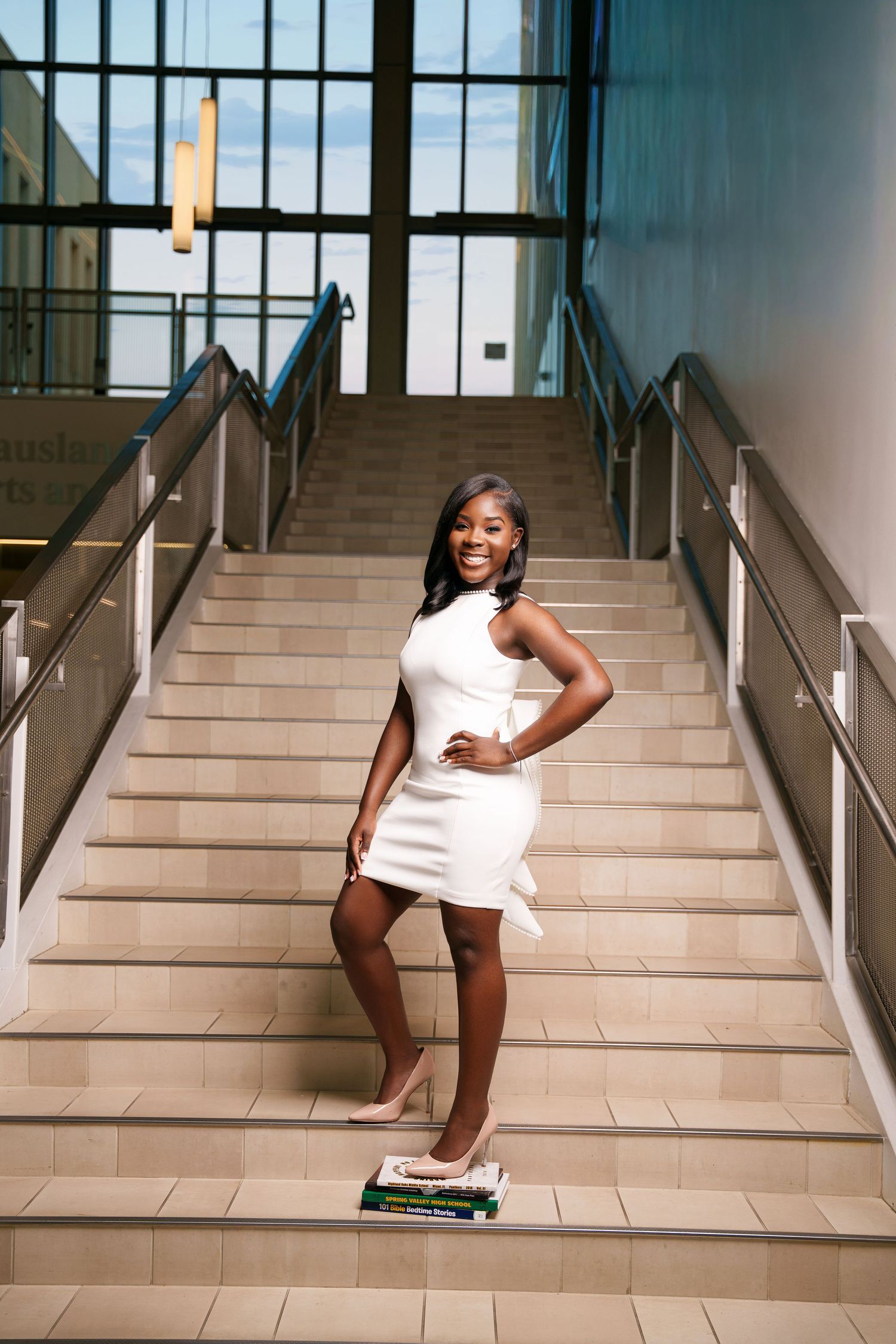Young woman in white dress posing confidently on indoor staircase with books, modern building interior.