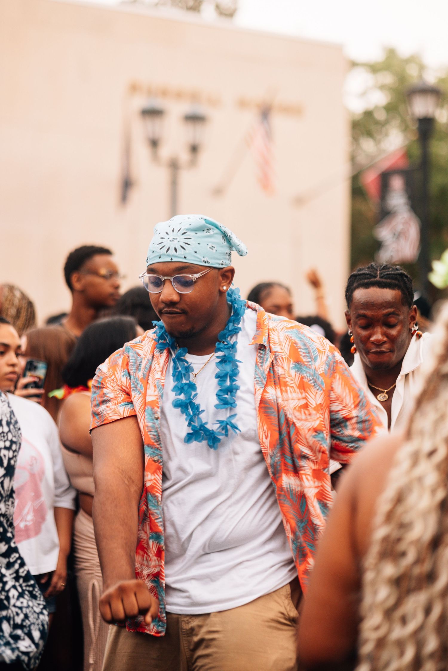 Person wearing a tropical Hawaiian shirt and blue bandana celebrates at an outdoor summer festival gathering.