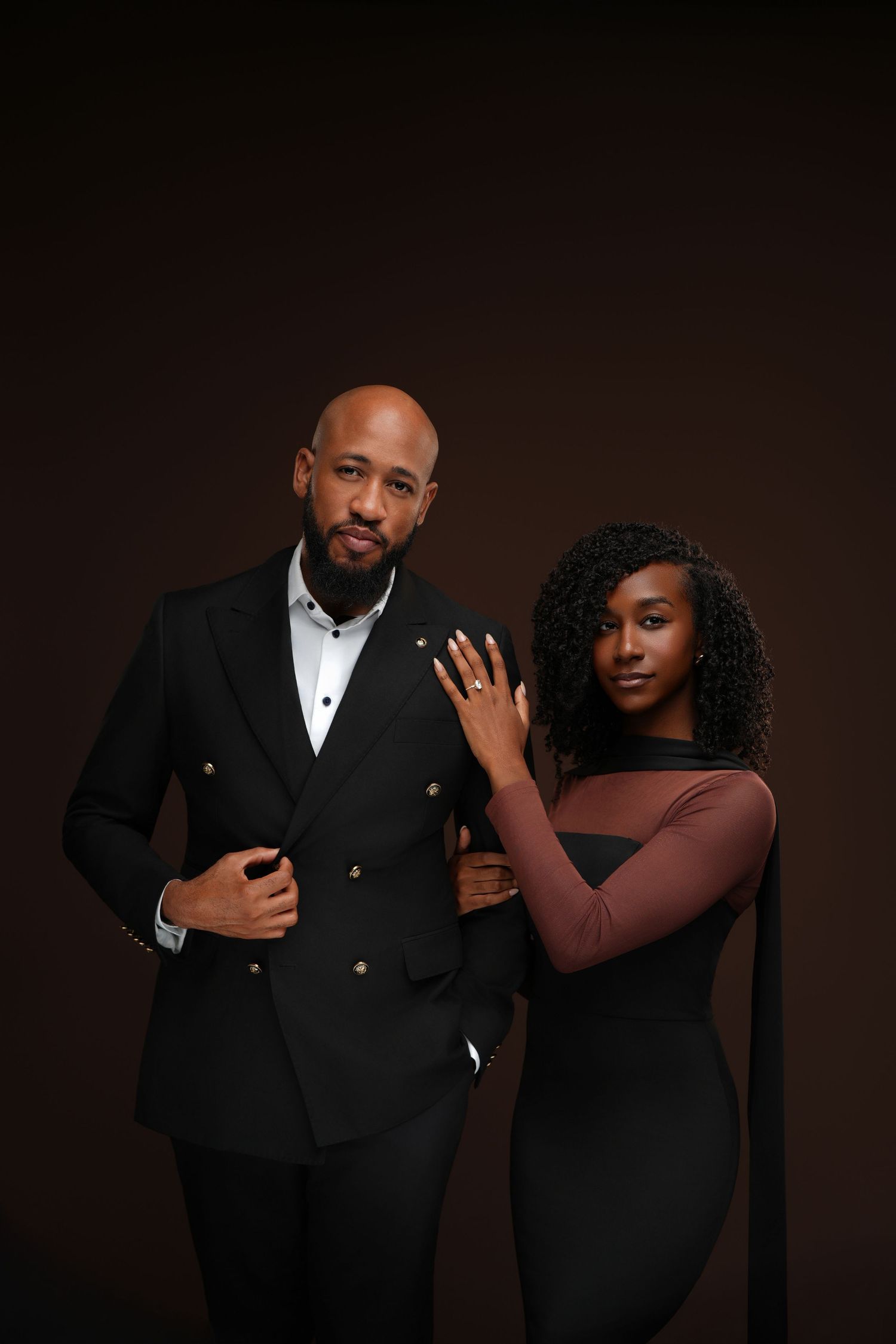 Elegant couple in black formal attire posing together against a dark brown studio background.