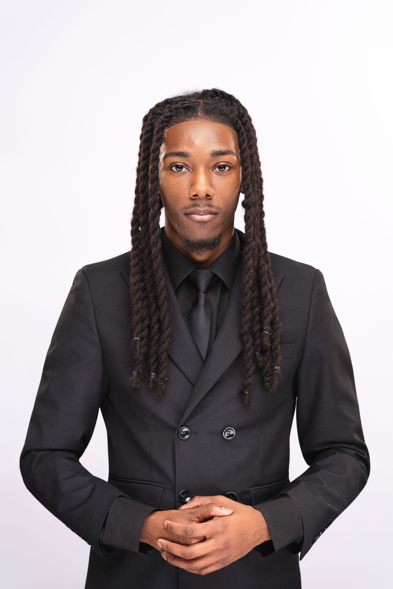 Young Black man with long dreadlocks wearing a black double-breasted suit and tie, posing against white background.