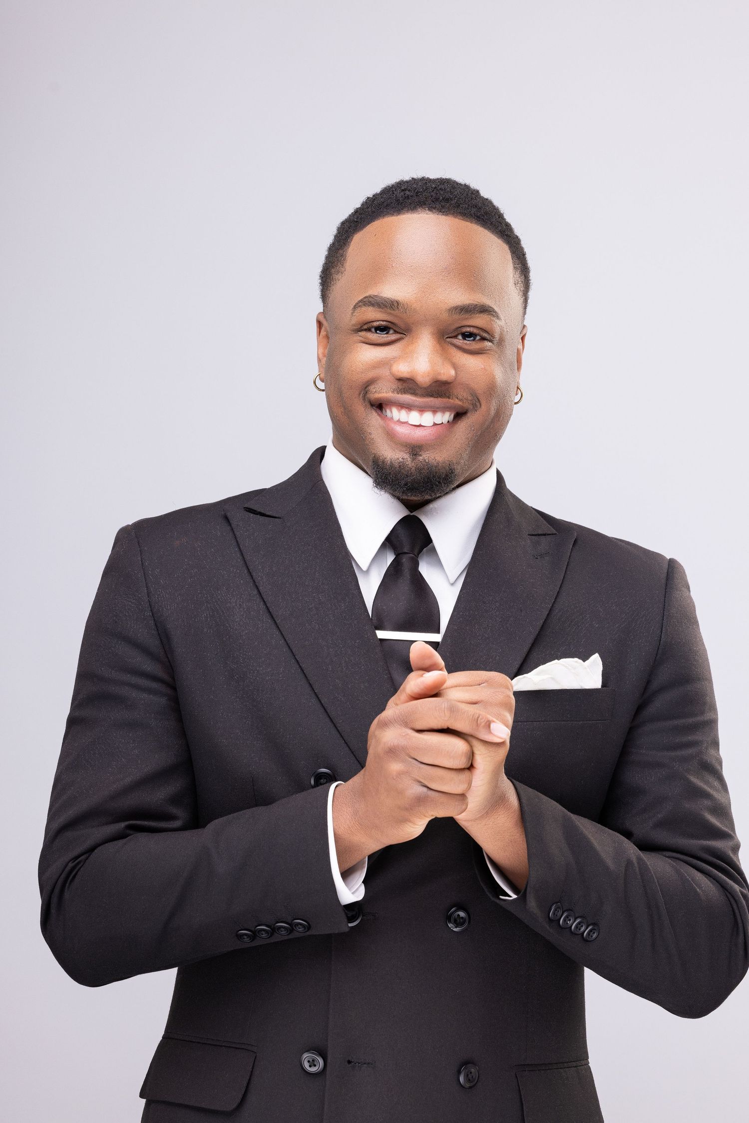 Person in black suit jacket smiling and gesturing with hands against white background.