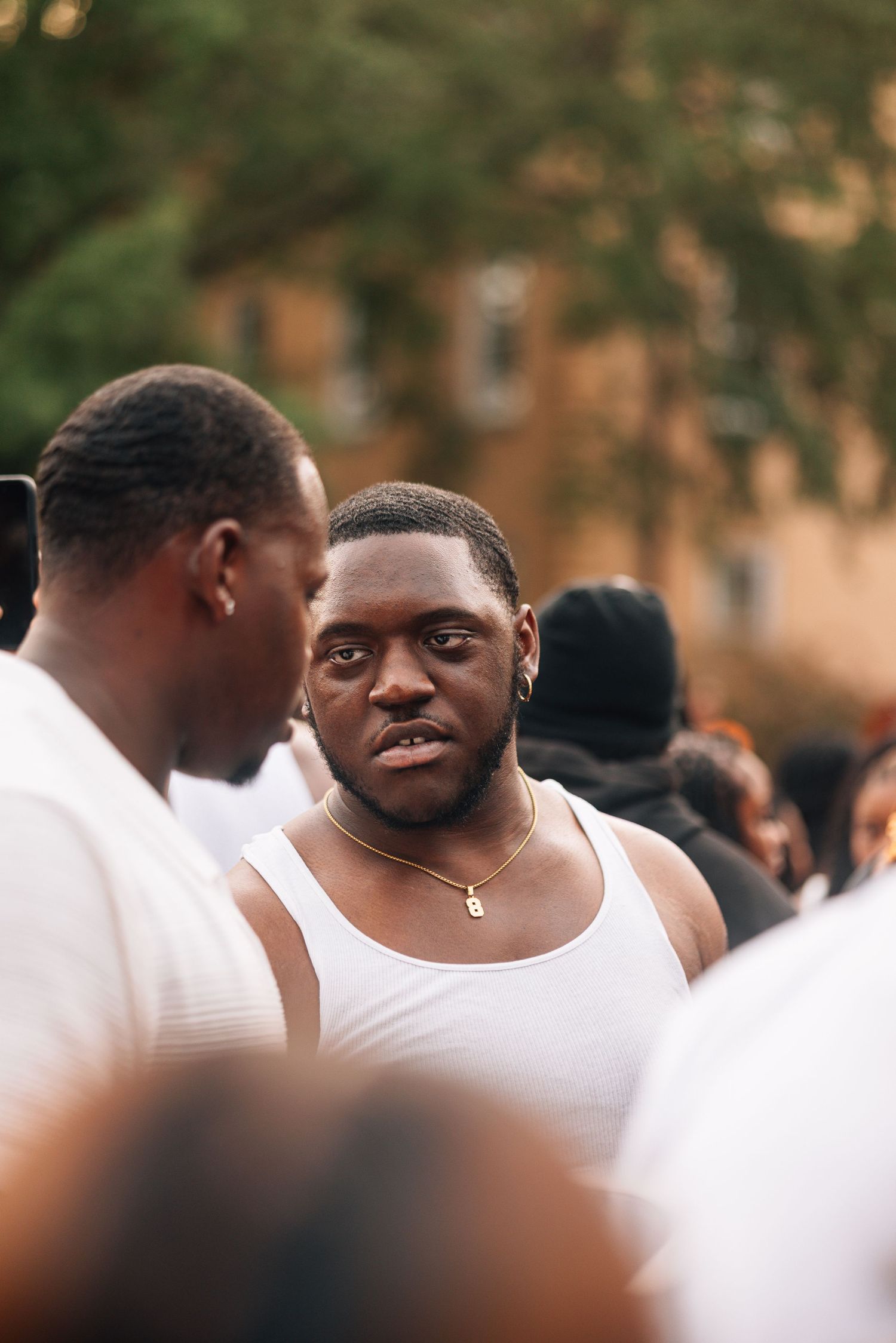 Two men in white tank tops having a conversation in a crowded outdoor setting during a warm day.