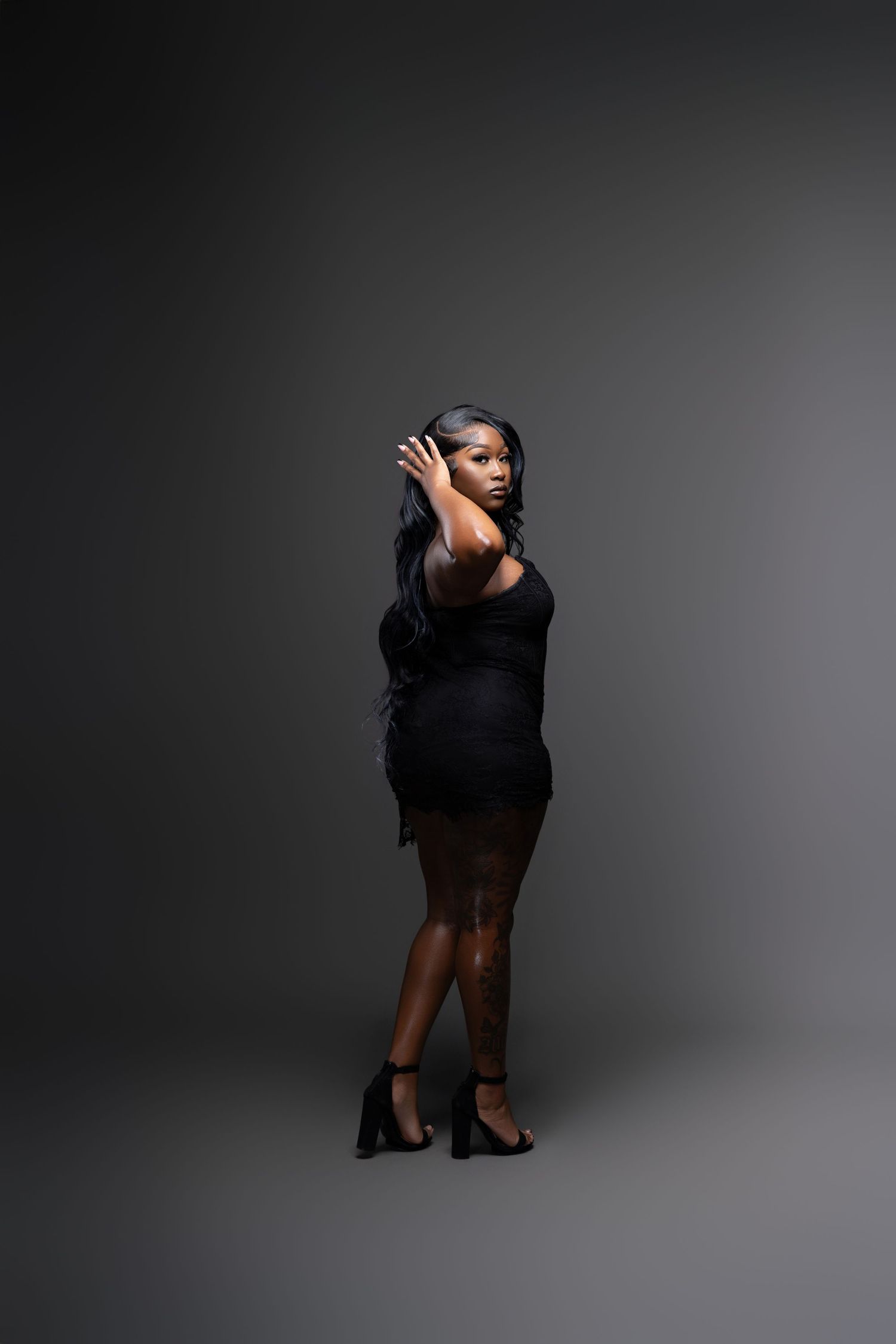 Fashionable portrait of a model wearing a black dress and heels against dark studio backdrop.