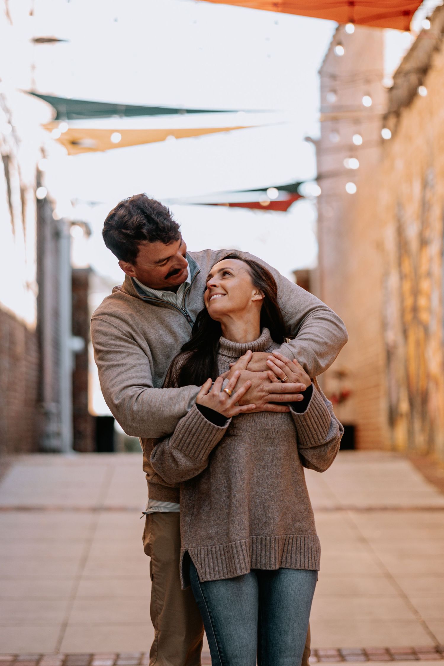 Couple embracing and laughing in an alleyway decorated with string lights and colorful fabric.