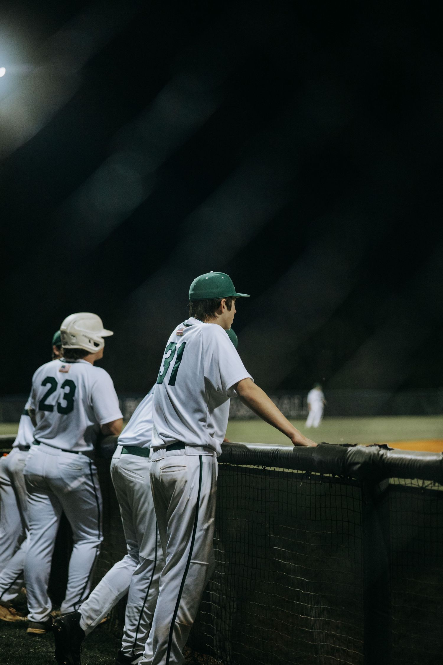Baseball players in white uniforms lean on the dugout fence watching the game.
