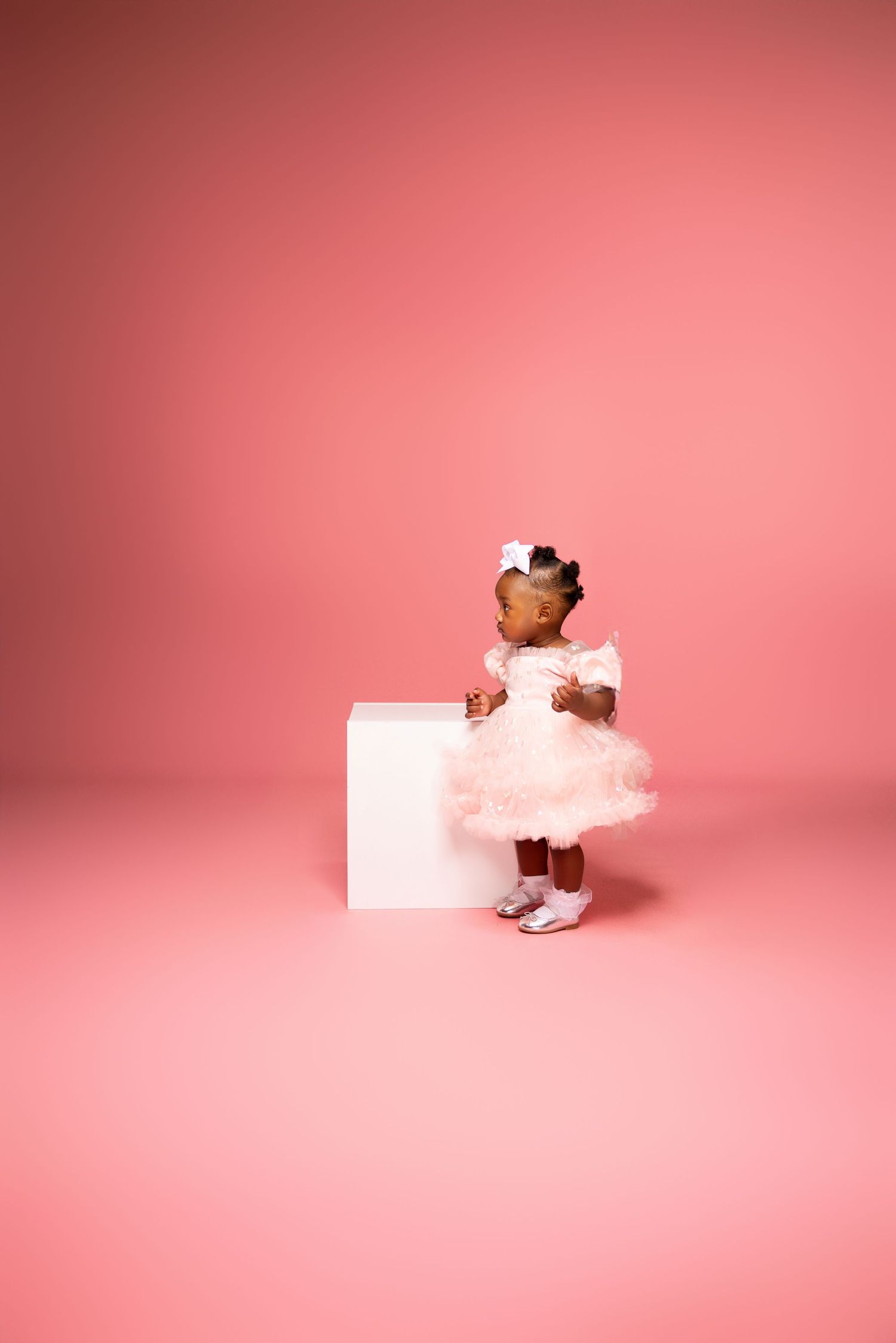 Small child in pink dress and bow standing next to white block against pink backdrop.