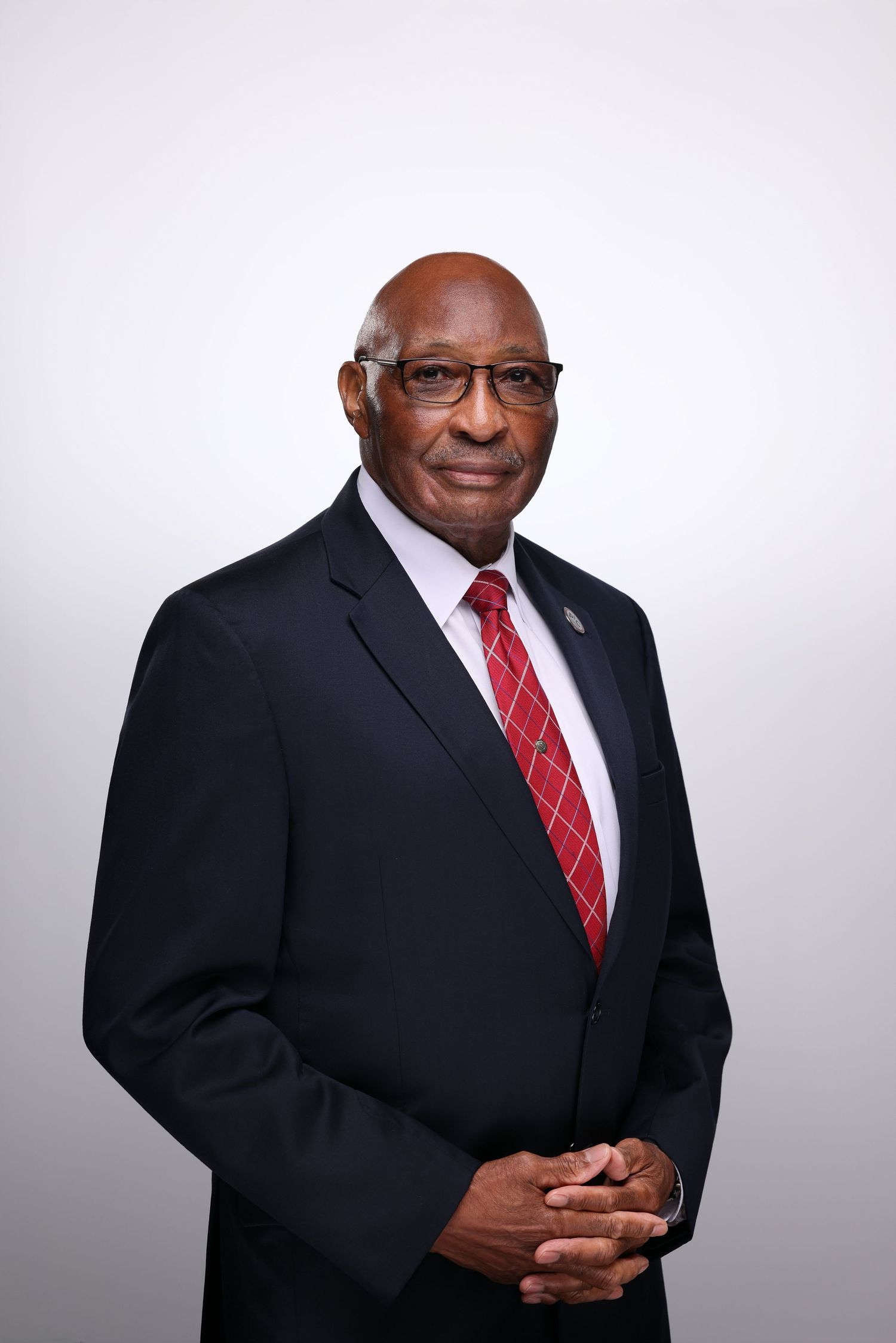 Professional headshot of a distinguished older man in a dark suit and red tie, wearing glasses, with hands clasped.