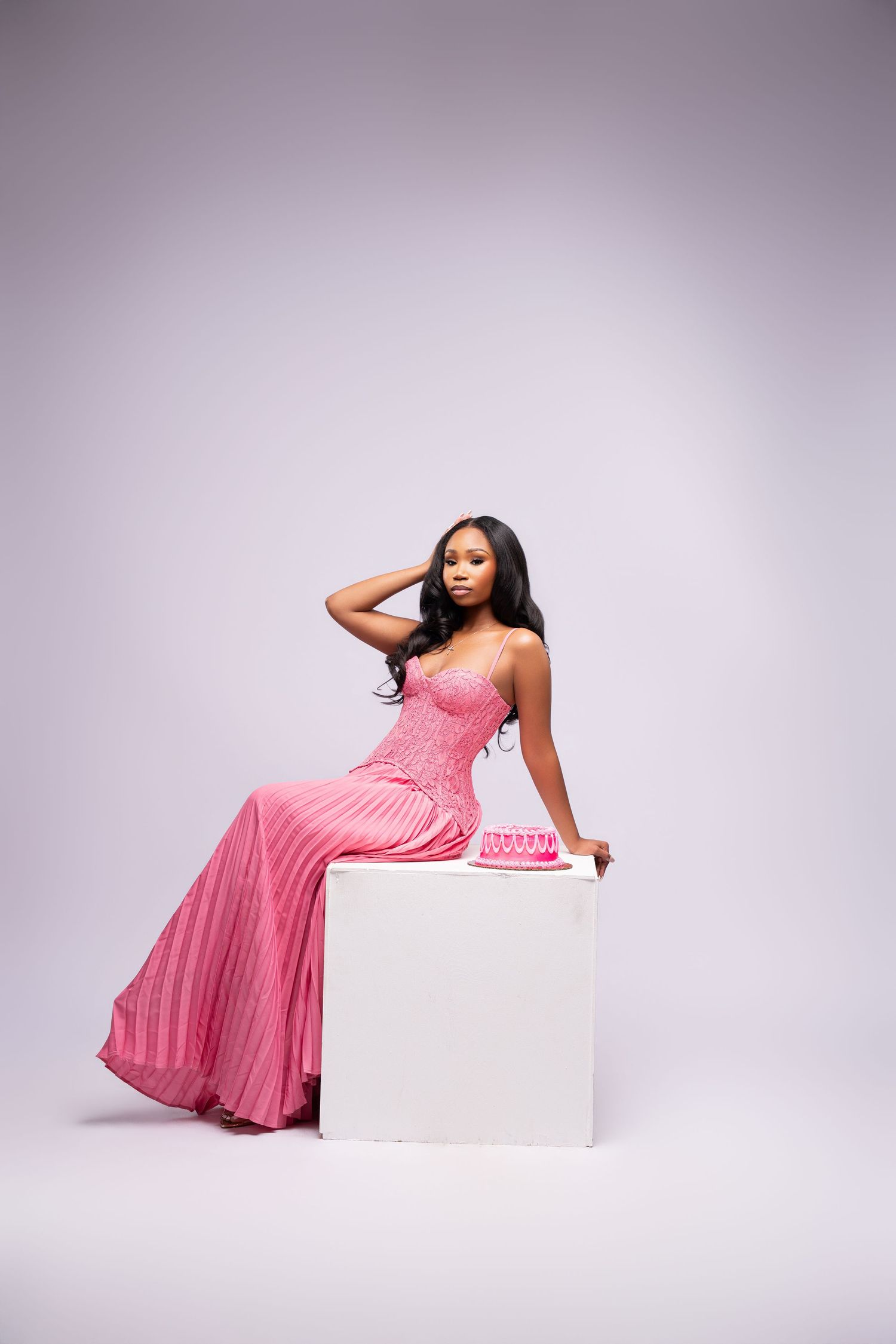 Woman in pink strapless pleated gown seated on white cube with matching pink clutch purse in studio.