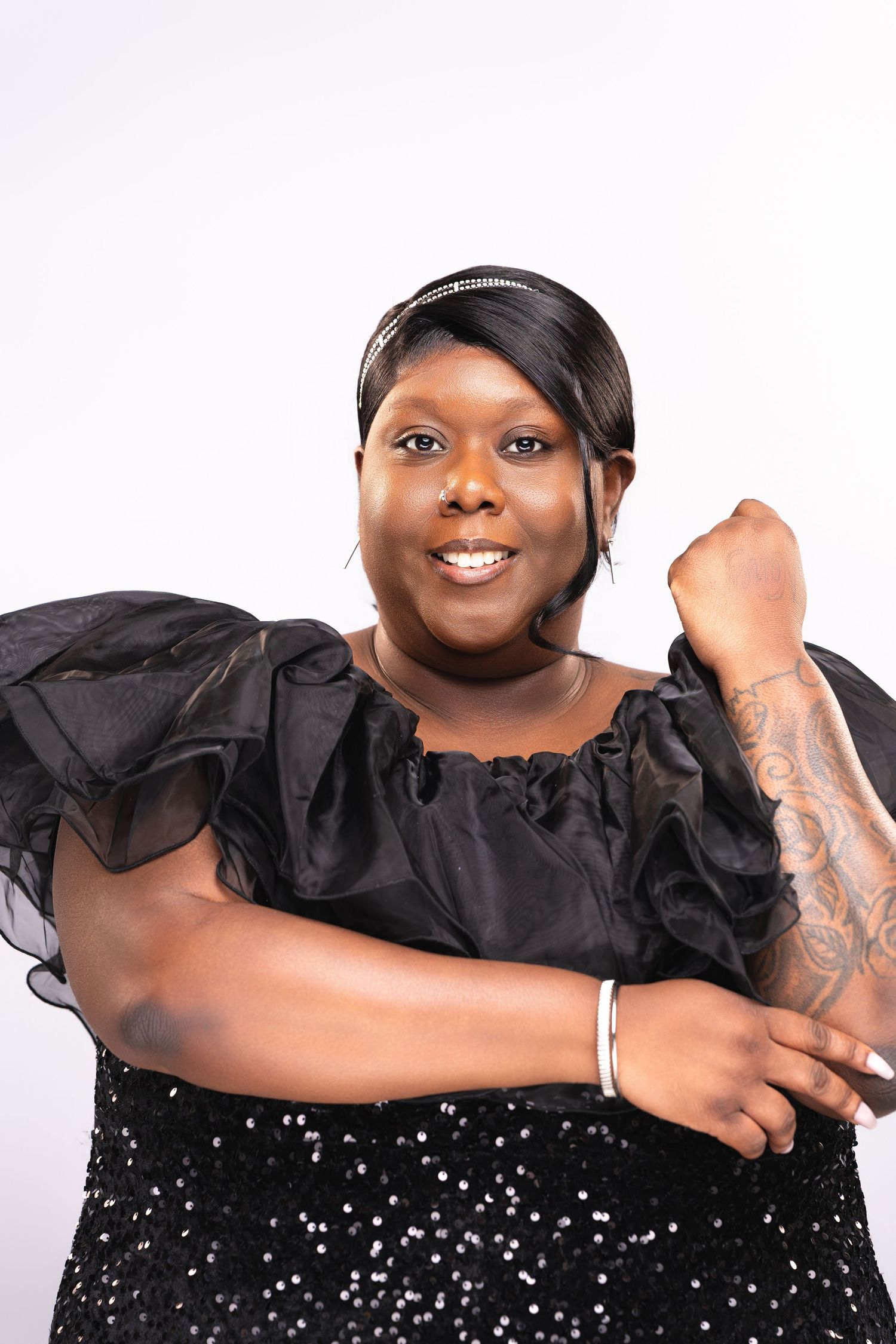Smiling woman in black ruffled sequin dress with tattoo, posing confidently against white background.