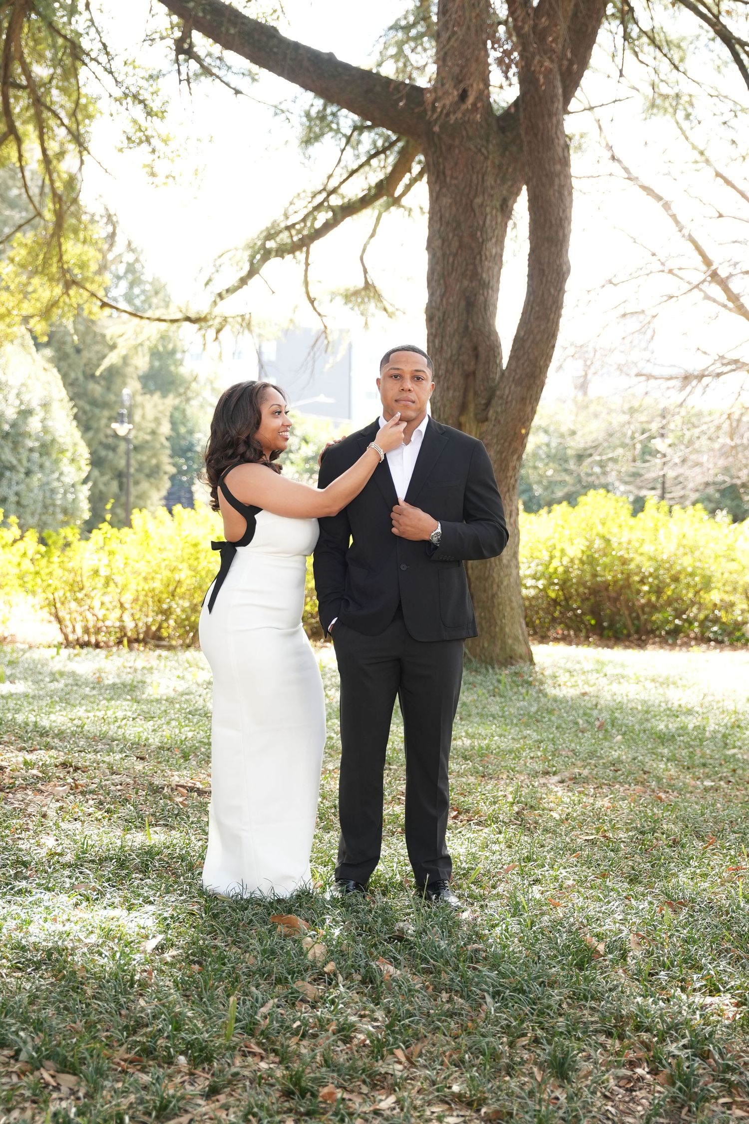 A couple in wedding attire share a romantic moment under a tree in a sunlit outdoor setting.