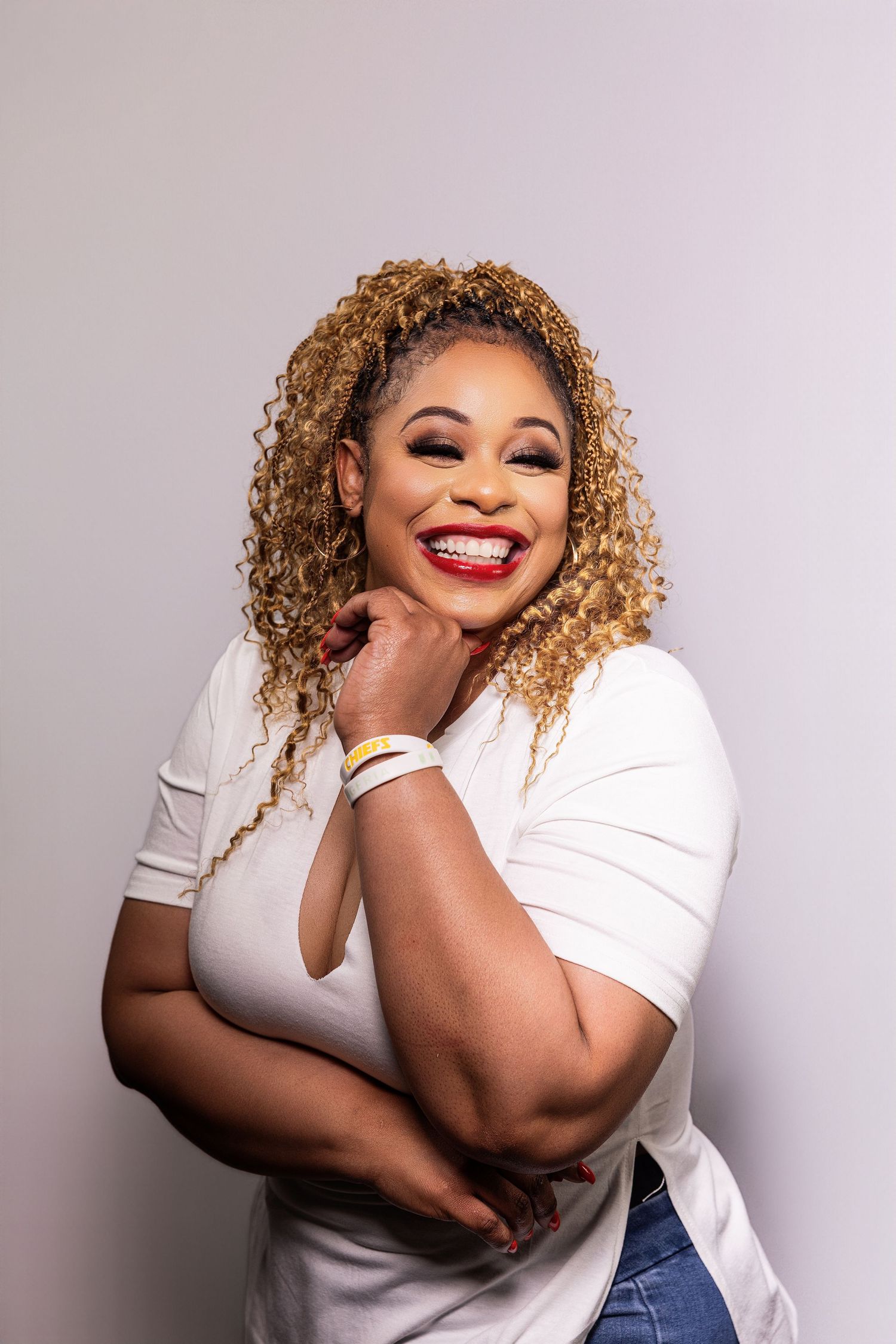 A joyful woman in a white shirt poses expressively against a light gray background with curly blonde hair.