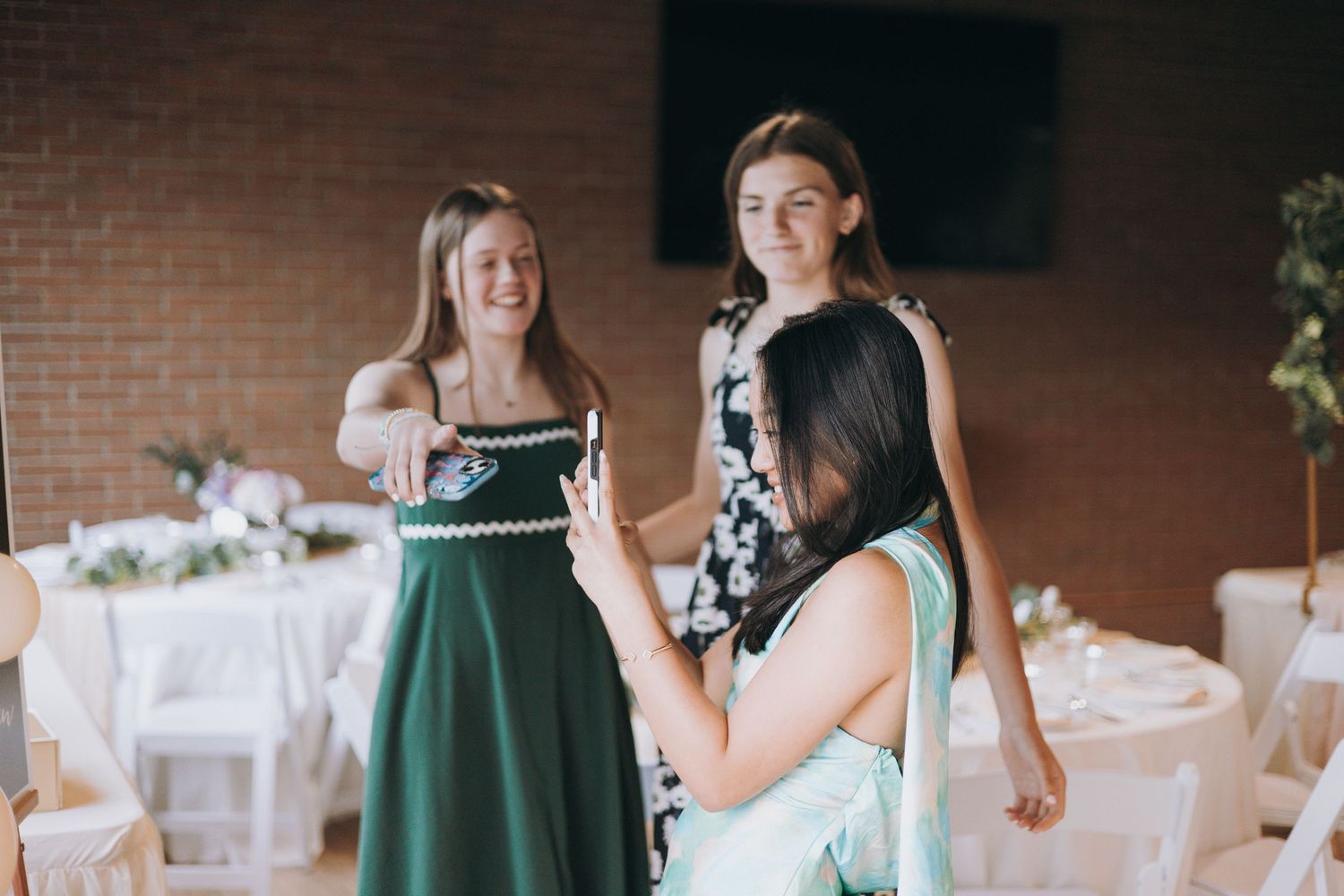 Three women fitting a green bridesmaid dress at an elegant wedding venue with white chair covers.