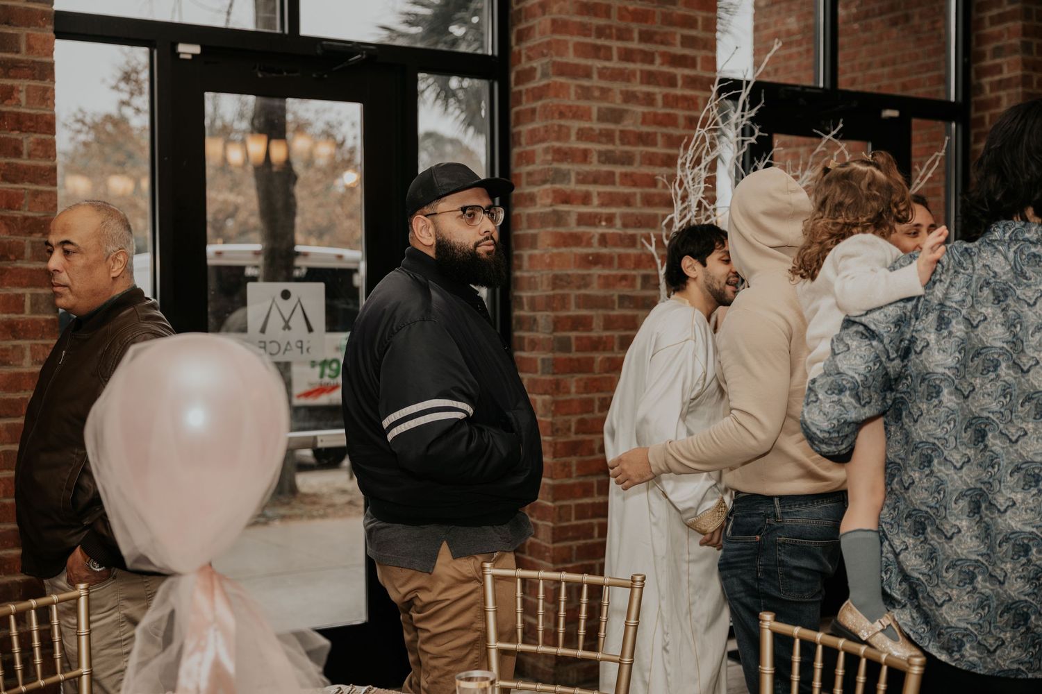 Guests mingling at an indoor event with gold chairs, pink balloons, and brick walls in a modern venue.