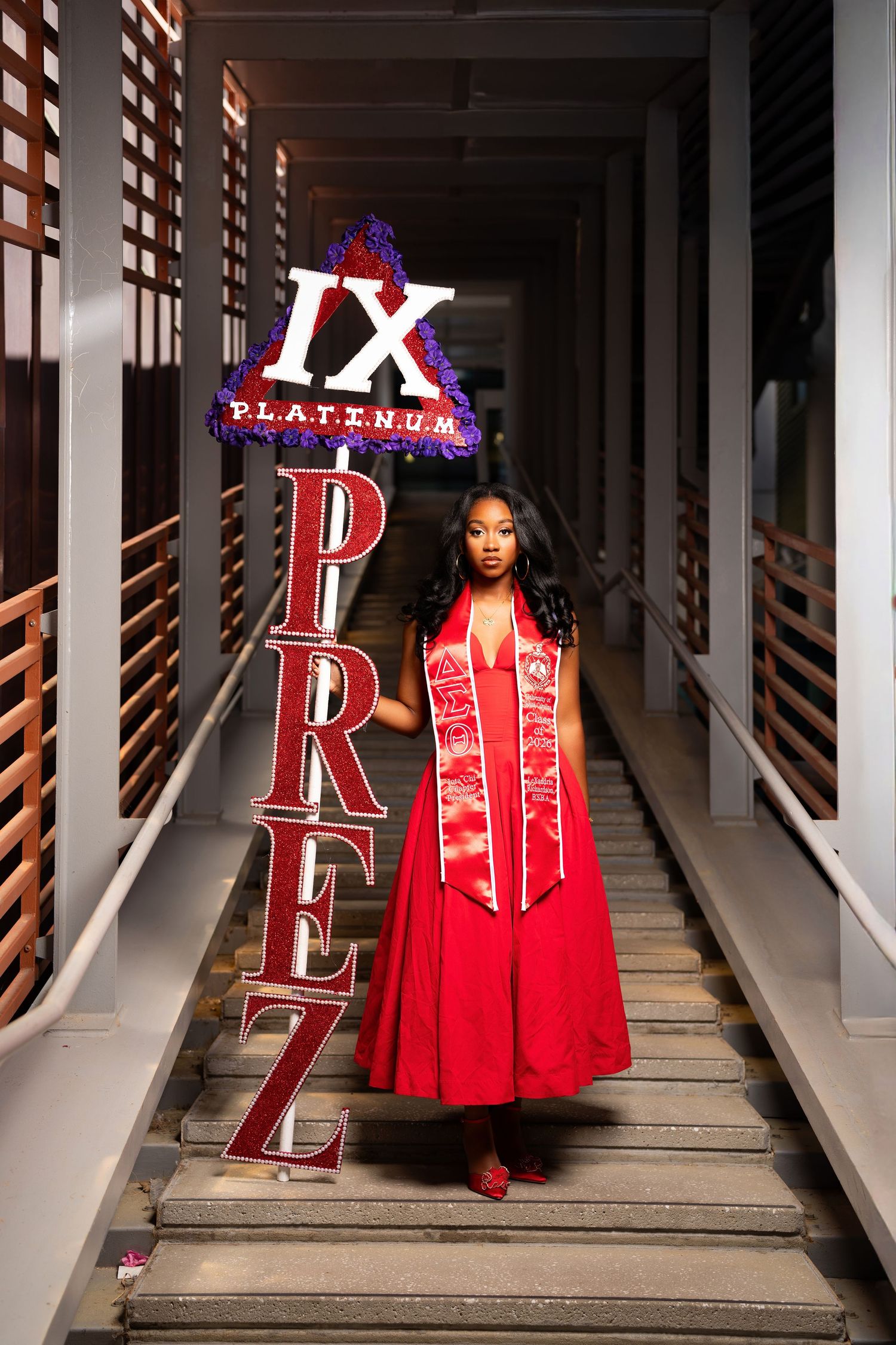 Woman in red graduation gown holding decorative letters spelling PREZ on stairs with IX Platinum sign.