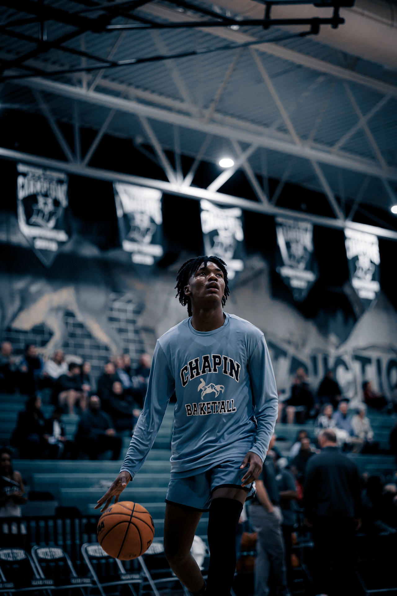 Basketball player wearing a light blue Chapin sweatshirt dribbles on a court under championship banners.