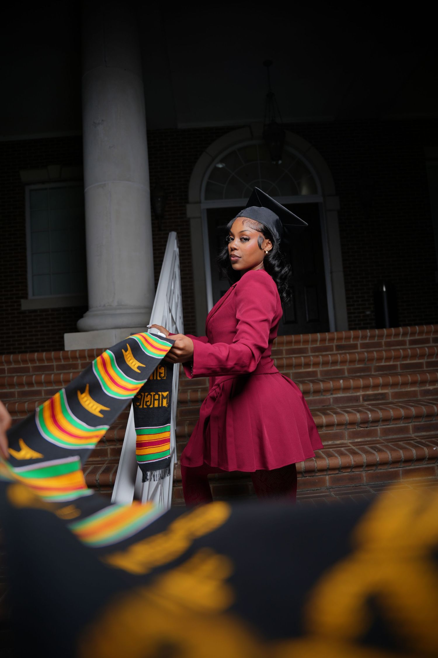 A person in a burgundy coat walks past colorful striped chairs in an outdoor setting with brick columns.