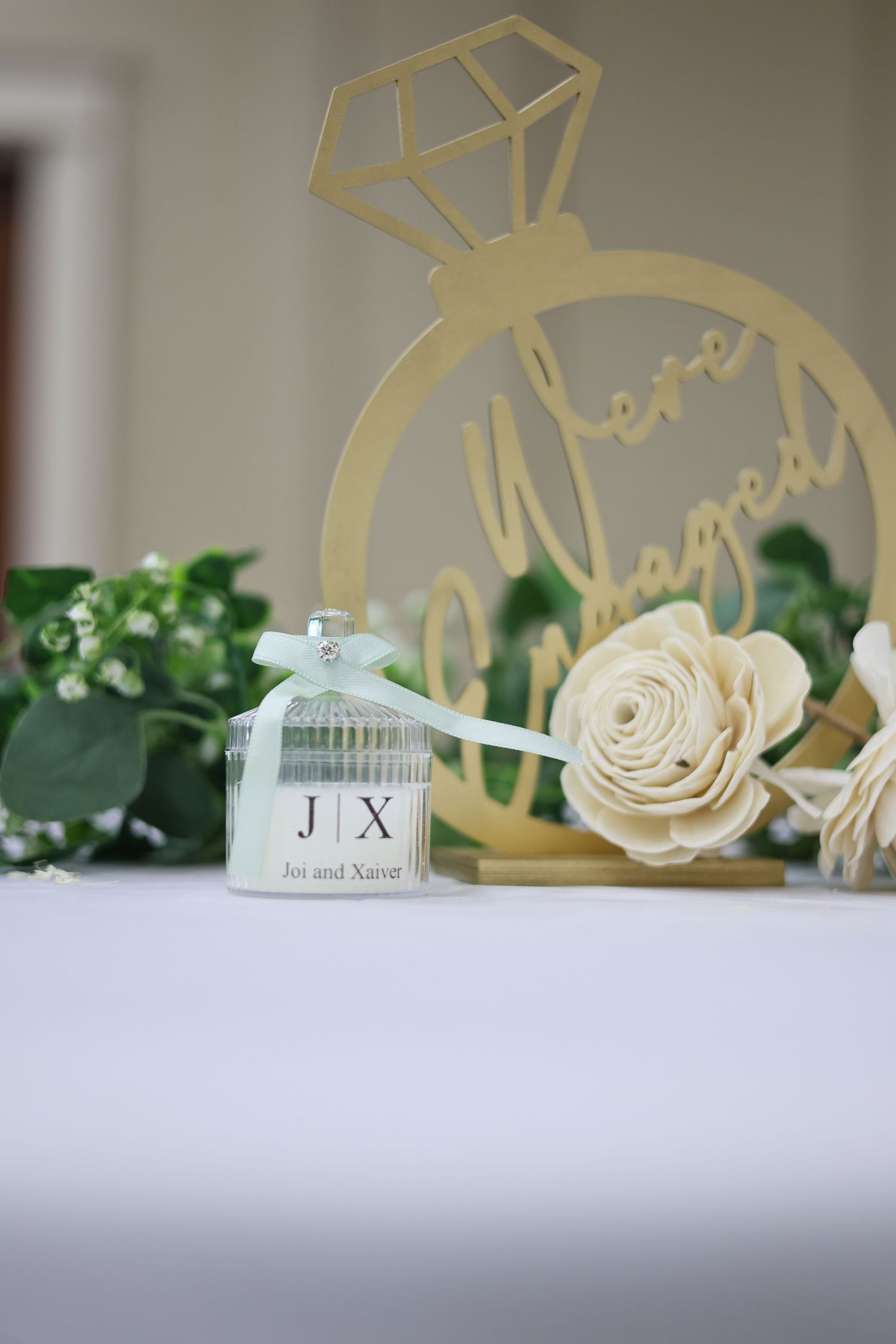 Gold engagement ring sign with 'We're Engaged' beside a personalized JX candle favor and white roses on a table.