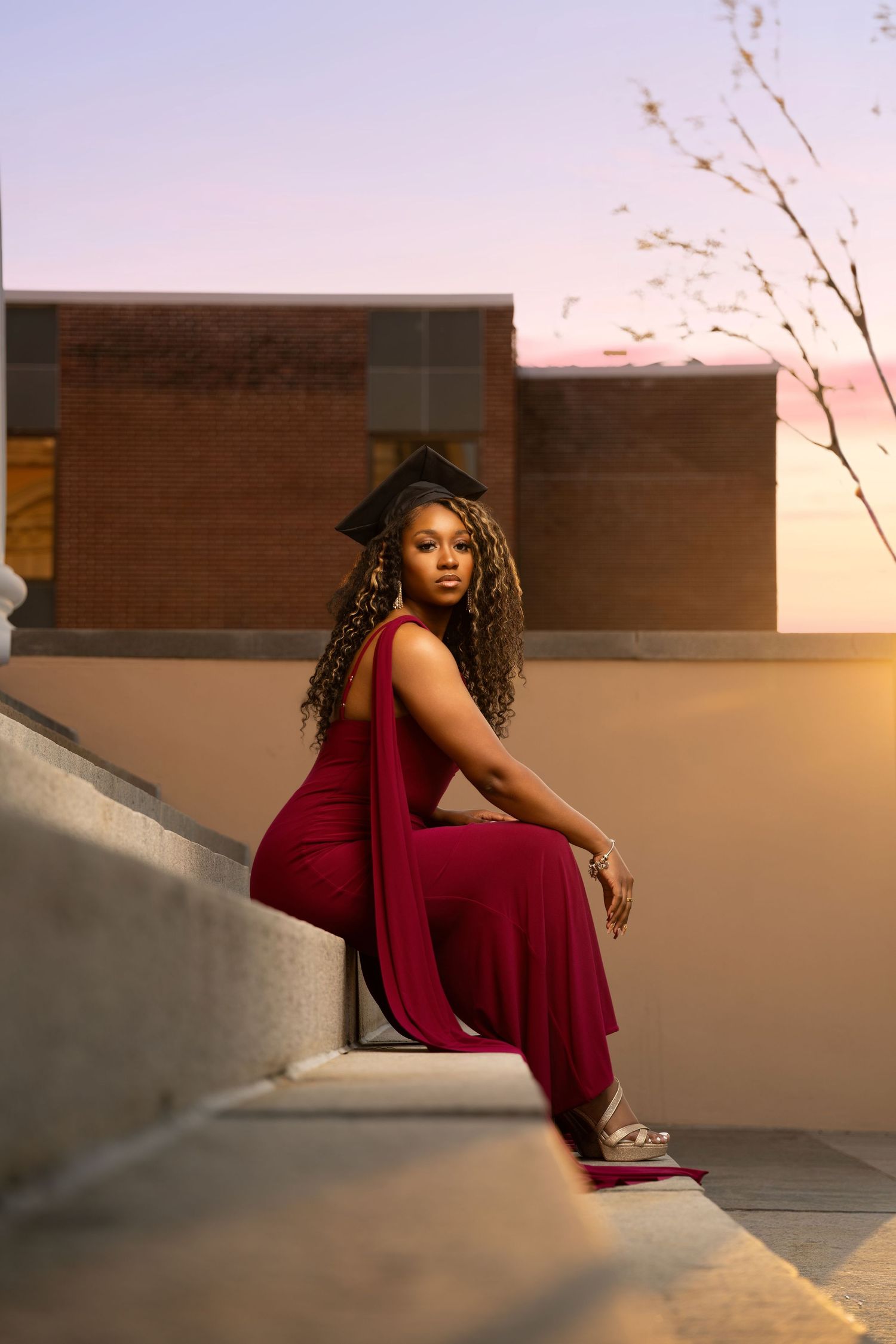 Young Black woman in red jumpsuit and graduation cap poses on steps at sunset.