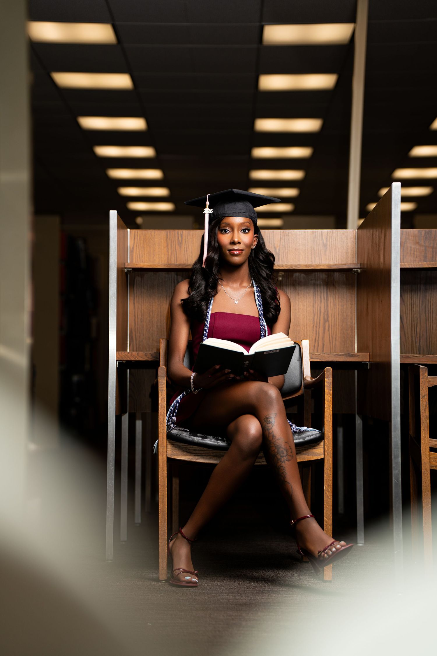 Young Black woman in graduation cap reads a book at a library study carrel, wearing a maroon dress and heels.