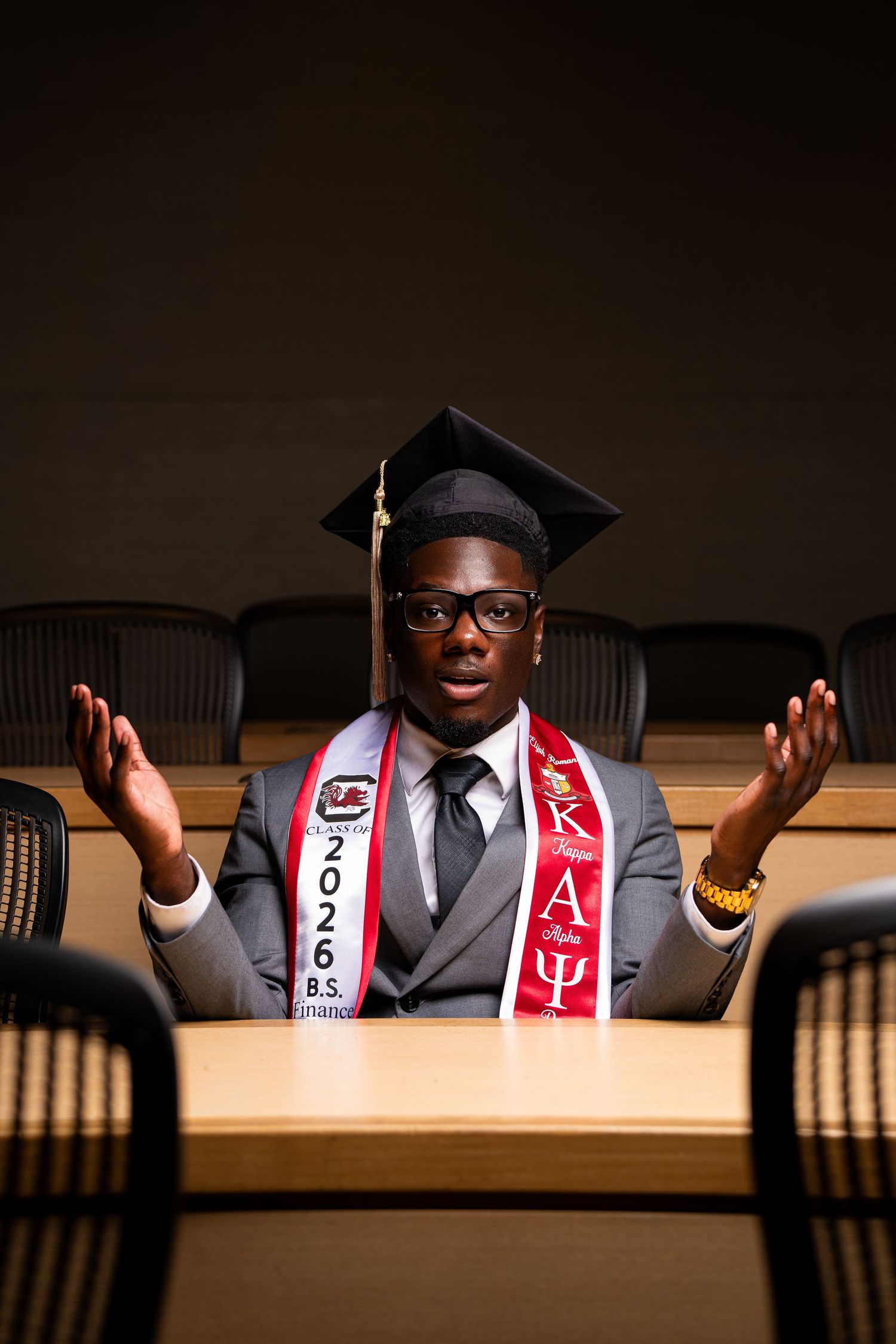 College graduate in cap and gown with 2026 B.S. sash and fraternity stole sits at conference table.