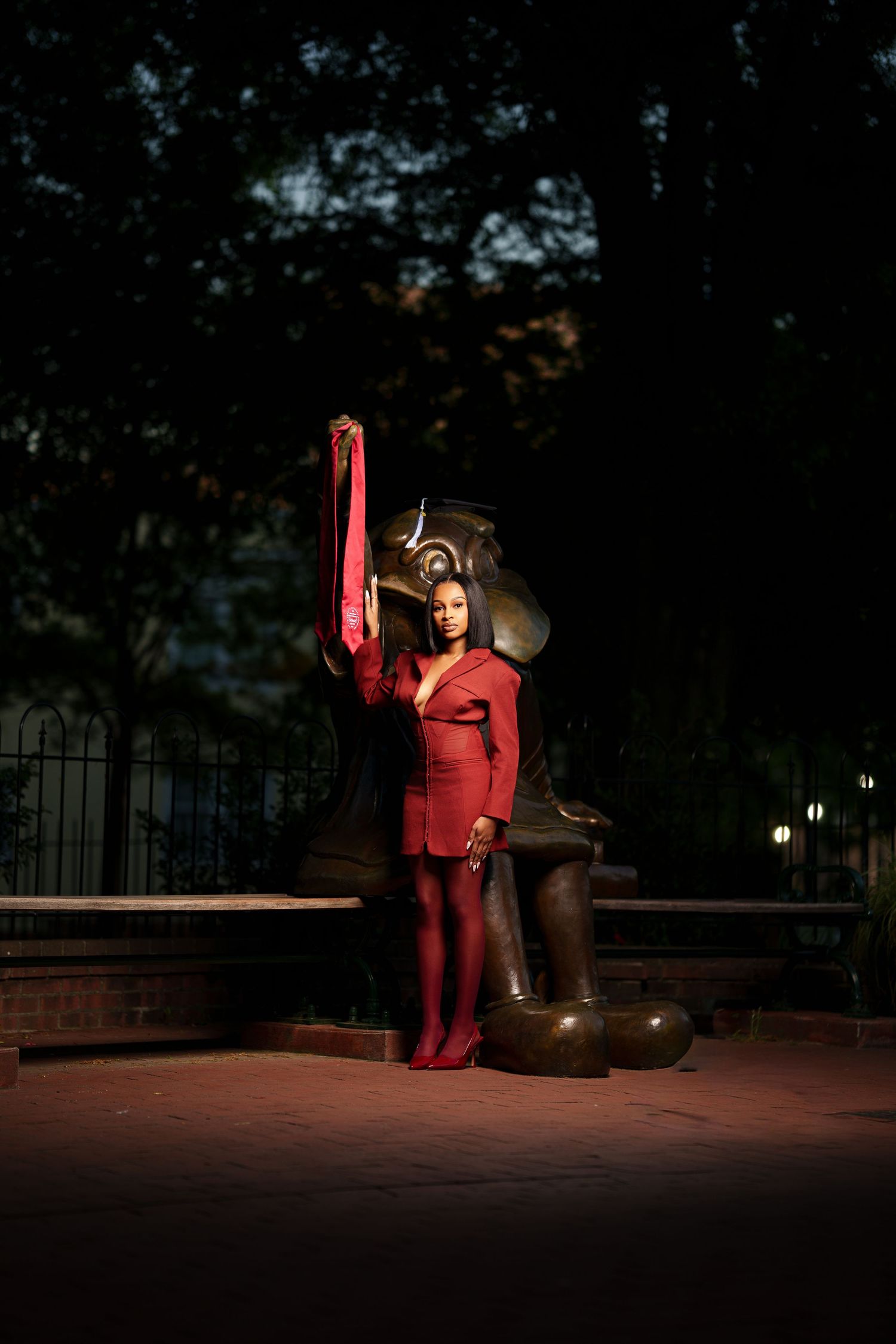 Woman in red dress holding red fabric beside bronze elephant statue at night.
