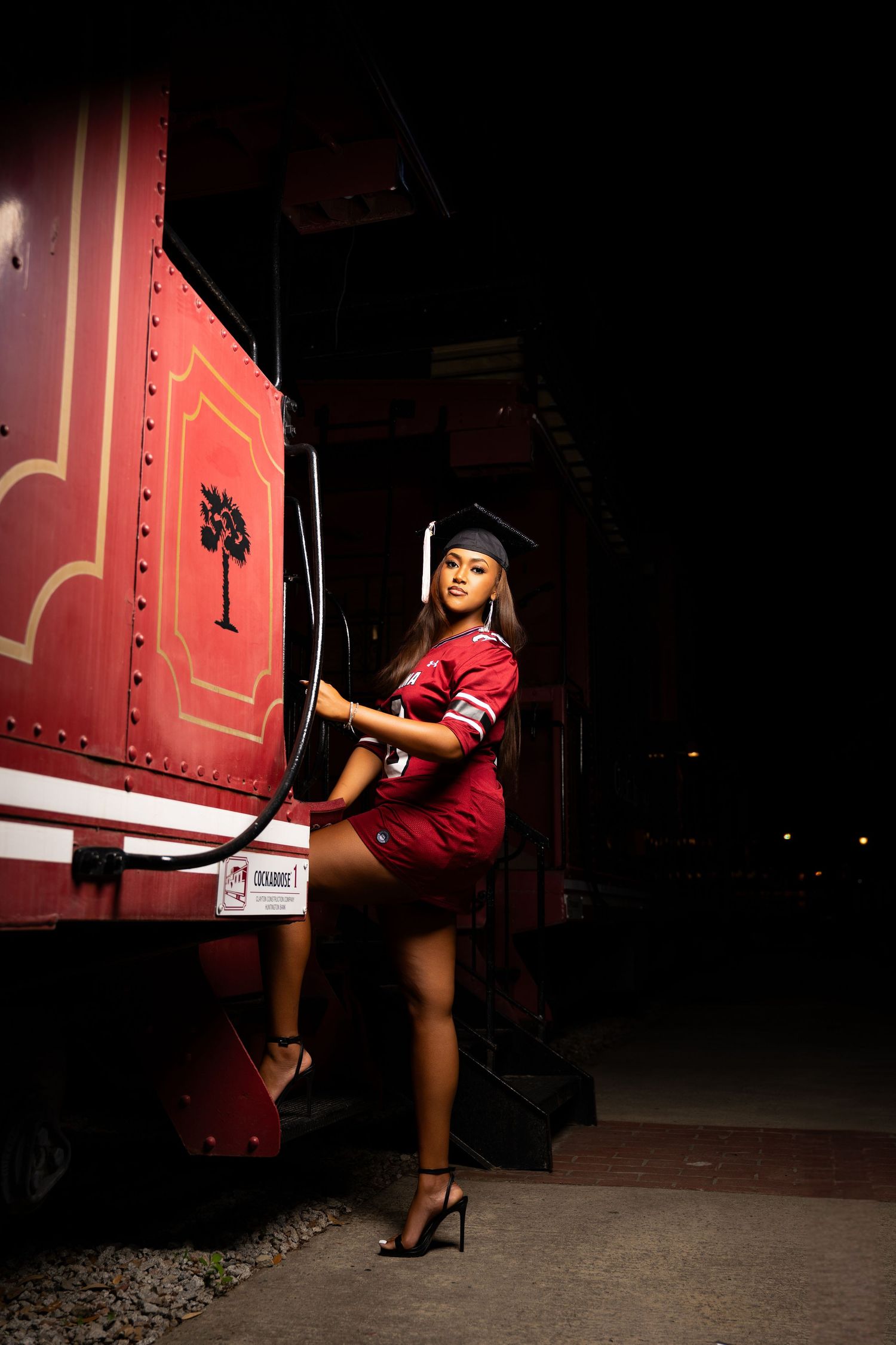 Young woman in red dress and graduation cap poses beside a vintage red train car at night.
